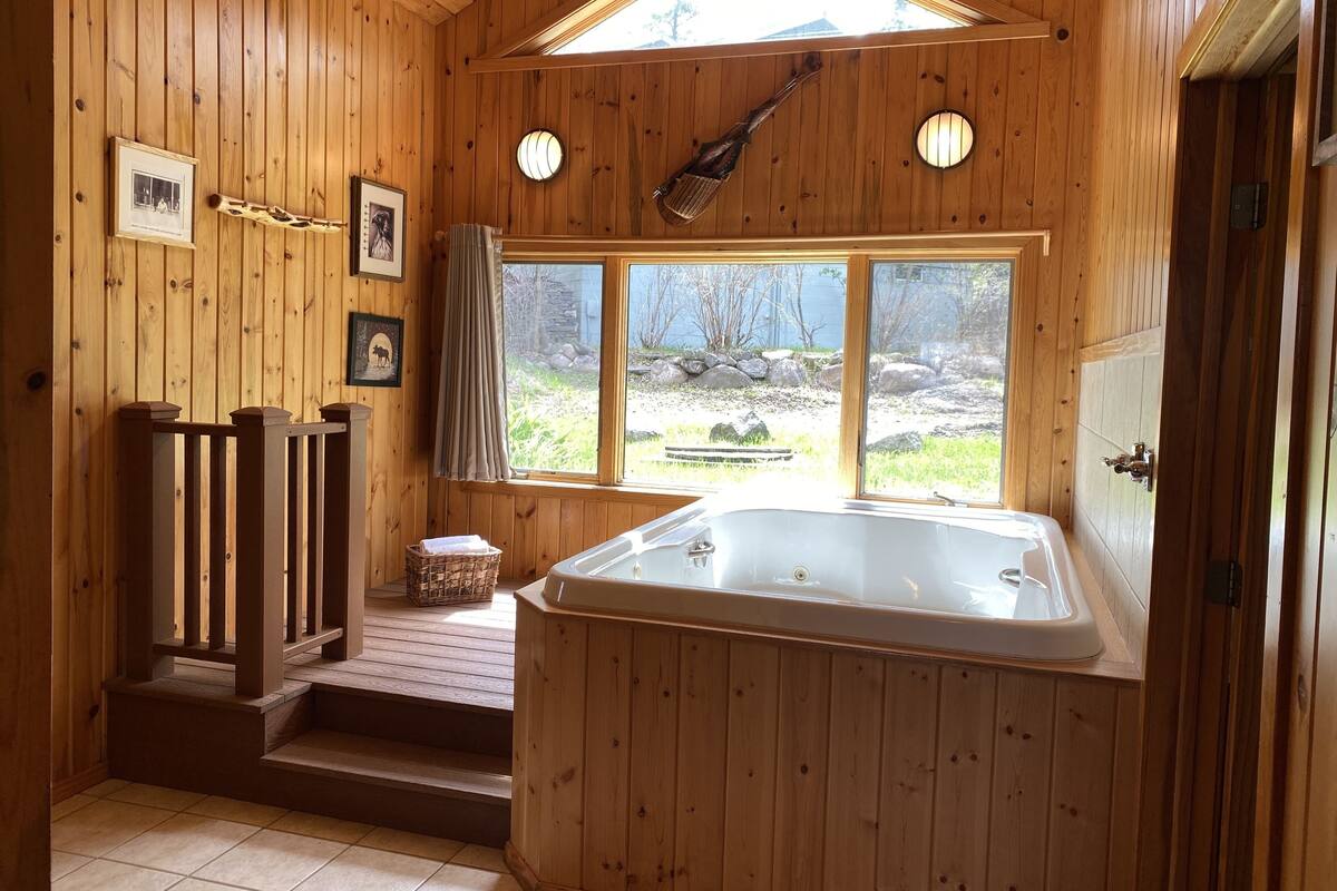 A wooden cabin bathroom with a large bathtub, wooden stairs, a window with a view of the outdoors, and framed pictures on the wall.