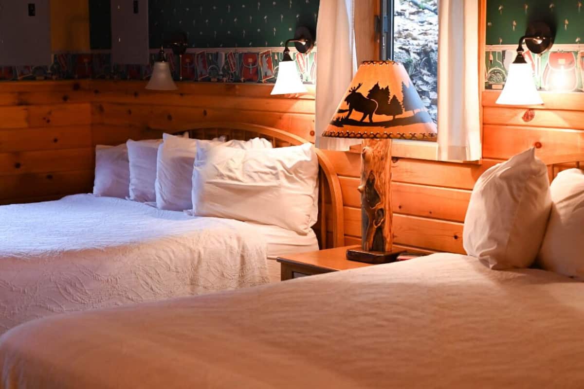 A cozy room with two beds, wooden walls, and a lamp with a moose design.