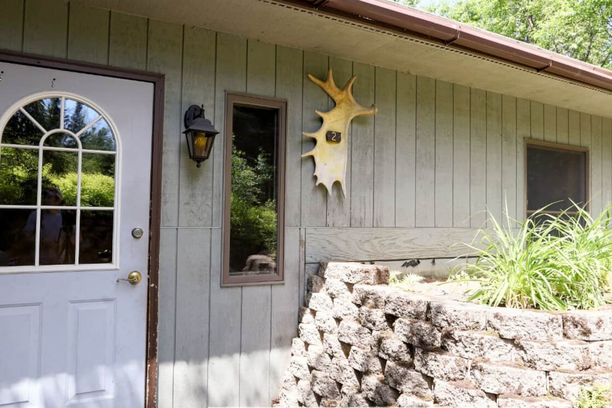 A rustic cabin with a white door, two windows, a stone wall, and a Native American animal head decoration.