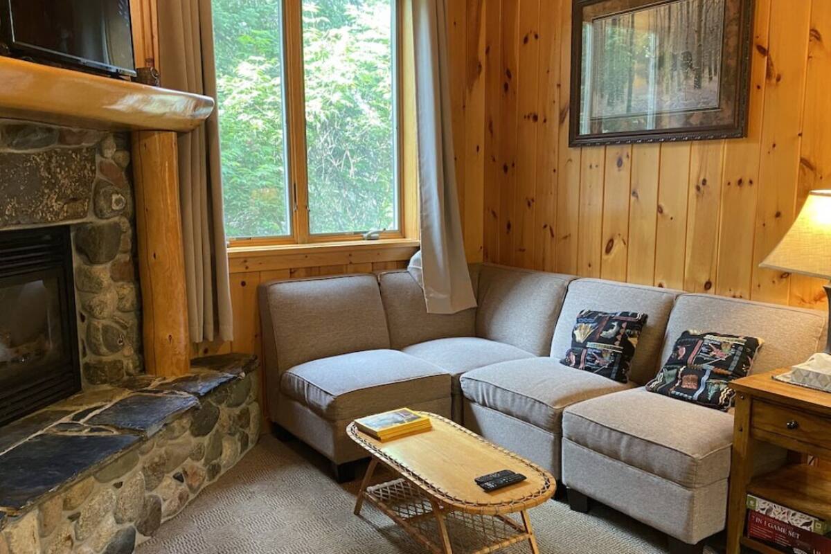 A cozy living room with a stone fireplace, a sofa set, a small table, and a window with a view of greenery.