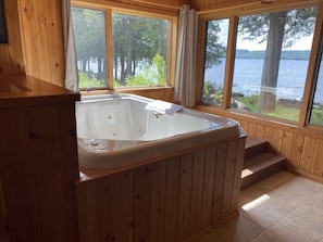 En stuga byggd av trä med ett bubbelbad, stora fönster och utsikt över en sjö.