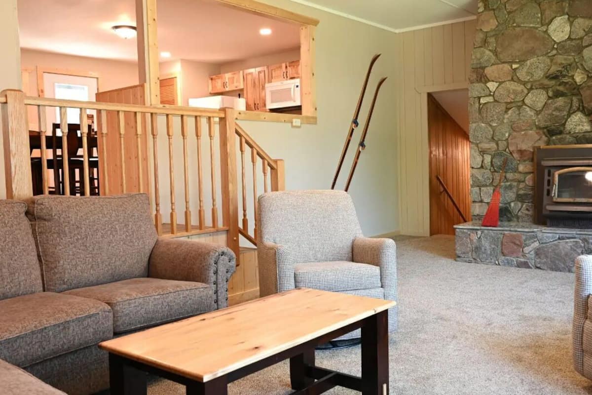 A cozy living room with a stone fireplace, a wooden staircase, and a view of the kitchen.