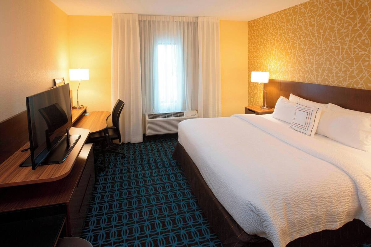 A hotel room with a bed, a desk with a computer, a chair, a window with curtains, and a patterned carpet.
