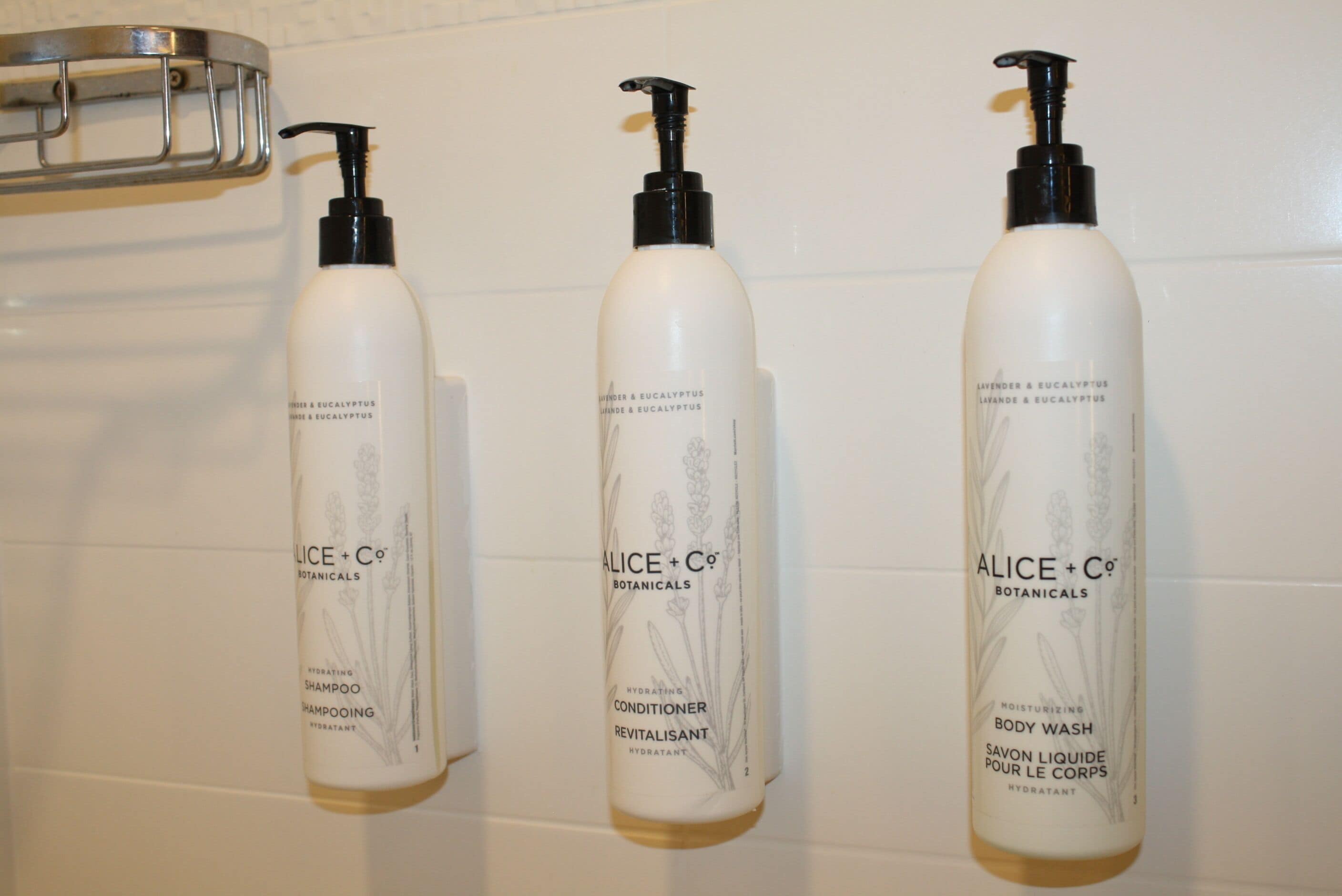 Three bottles of Alice + Co Botanicals body wash and conditioner on a white tiled wall.