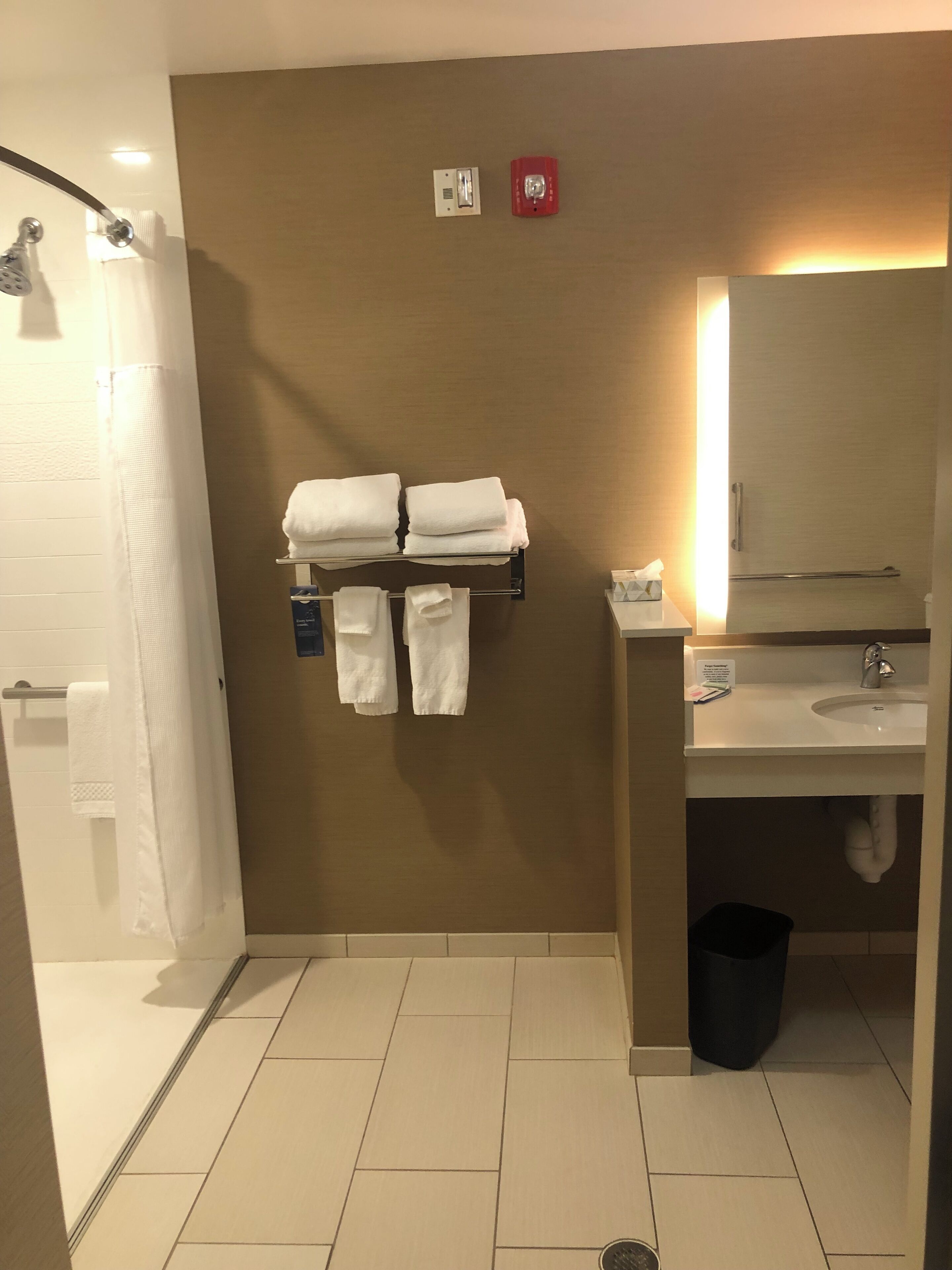 Un baño con ducha, lavabo y espejo. Hay toallas colgadas en un perchero y un cubo de basura en el suelo.