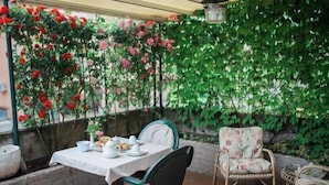 An outdoor patio with a table set for dining, surrounded by wicker chairs and a backdrop of climbing roses.