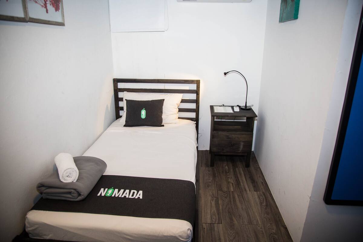 A small, neatly made bed with a wooden headboard, a nightstand with a lamp, and a wall-mounted TV.