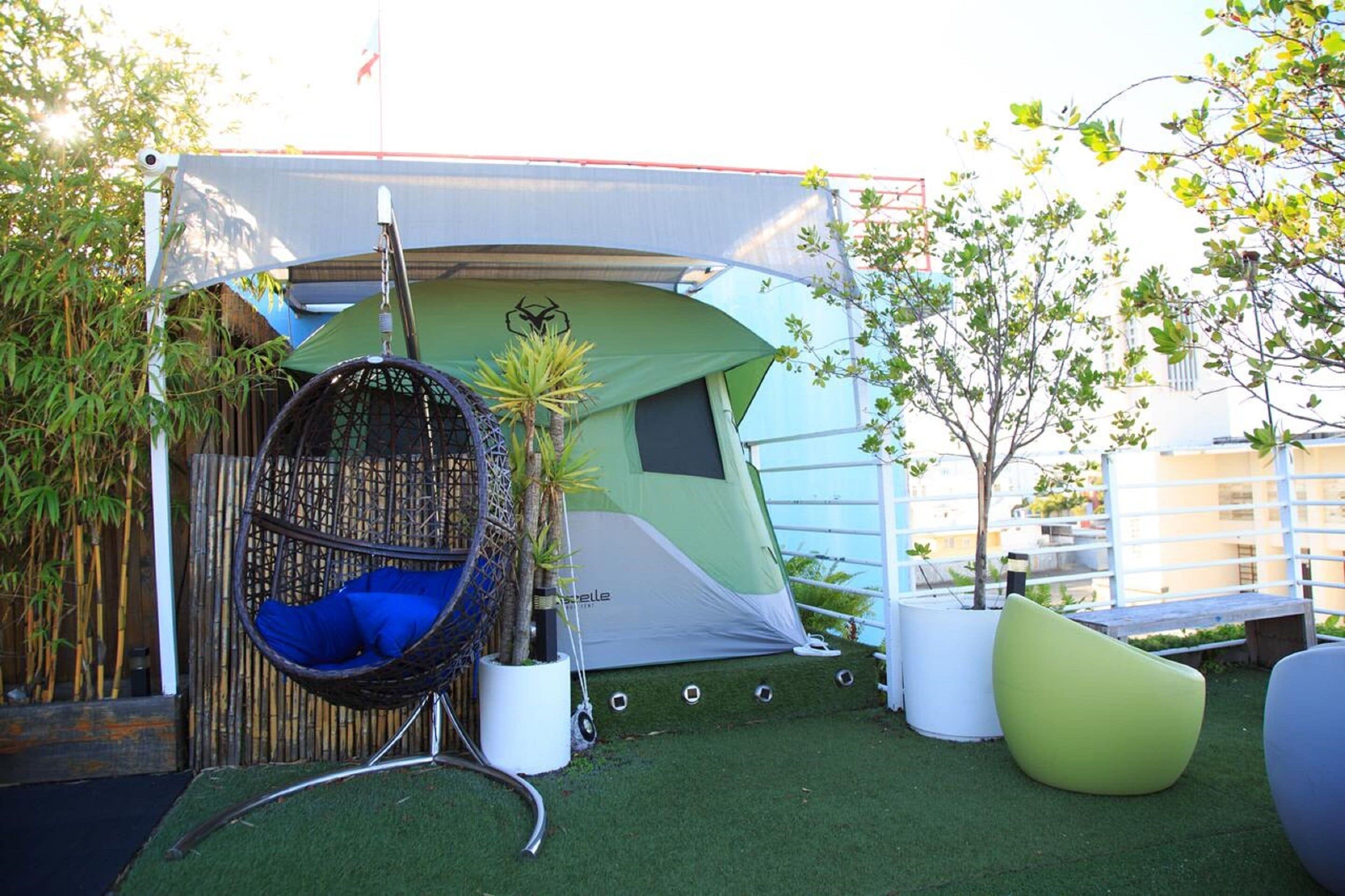Una carpa con diseño verde y blanco, un sillón colgante con cojín azul y una planta en maceta.