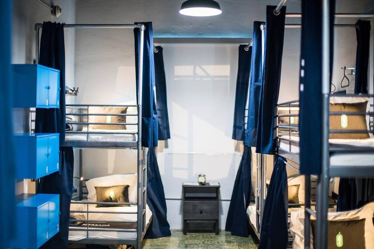A dormitory room with bunk beds, a small desk with a clock, and a window with curtains.
