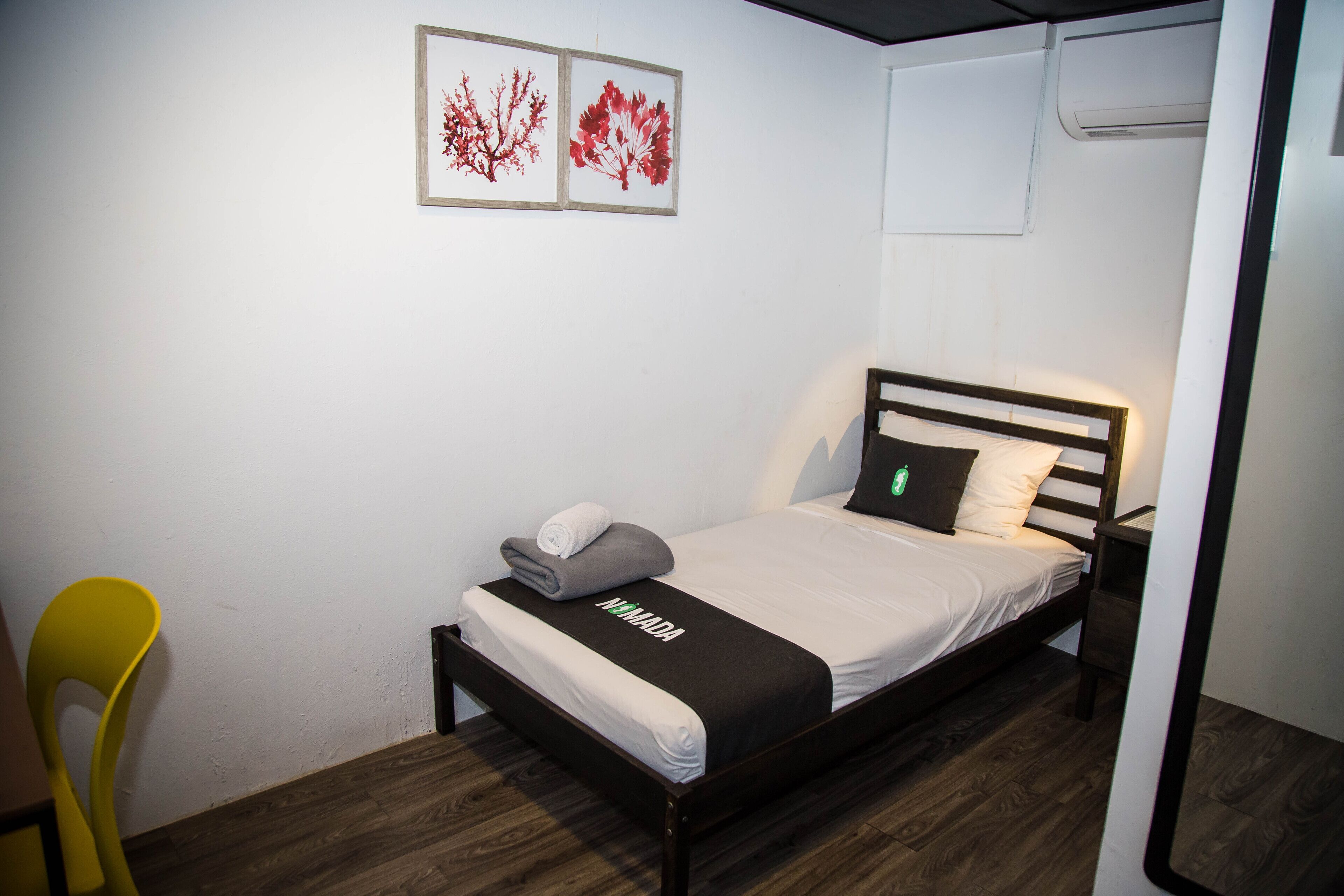 Un lit simple avec une tête de lit noire, un matelas blanc et une couverture noire ornée du mot « HEIMADA ». Il y a une chaise jaune sur la gauche et un climatiseur fixé au mur au-dessus du lit.