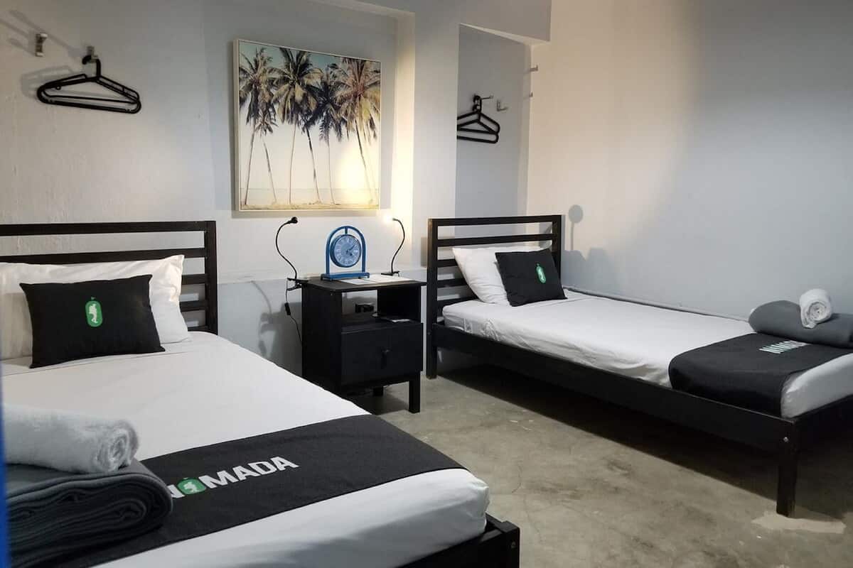 A hotel room with two beds, a nightstand, a clock, and a painting of palm trees.