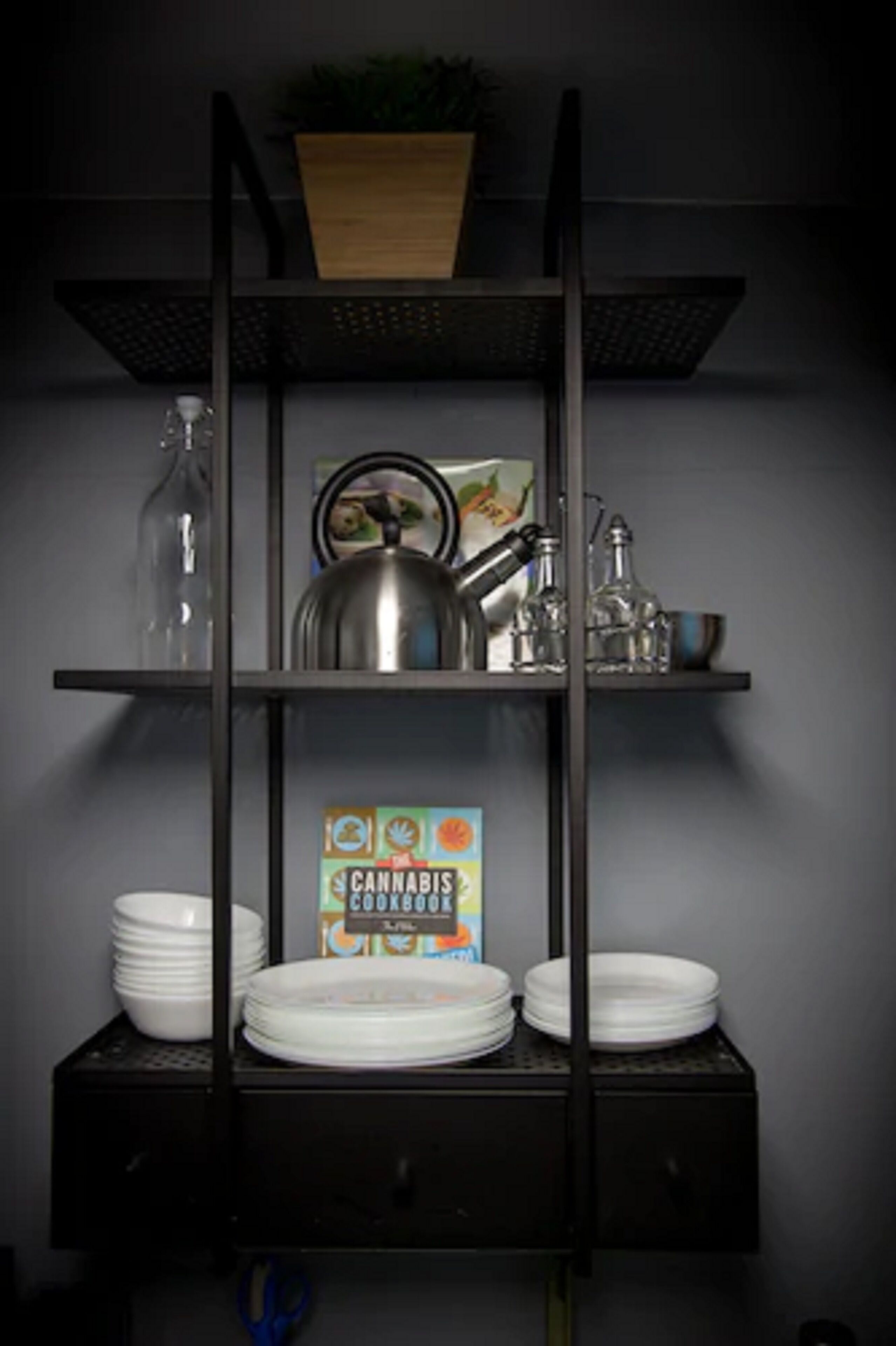 A kitchen shelf with a cannabis cookbook, plates, a kettle, and glassware.