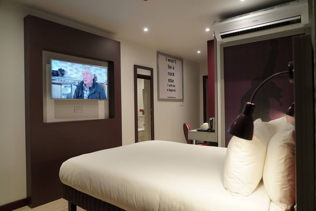 A hotel room with a large bed, a television mounted on the wall, a mirror, and a poster.