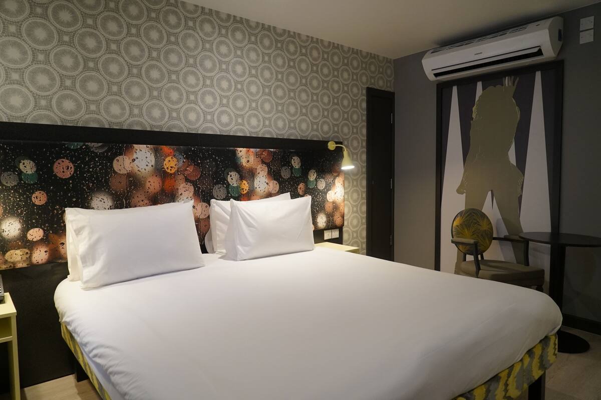 A hotel room with a bed, a wall with a circular pattern, an air conditioning unit, and a silhouette artwork on the wall.