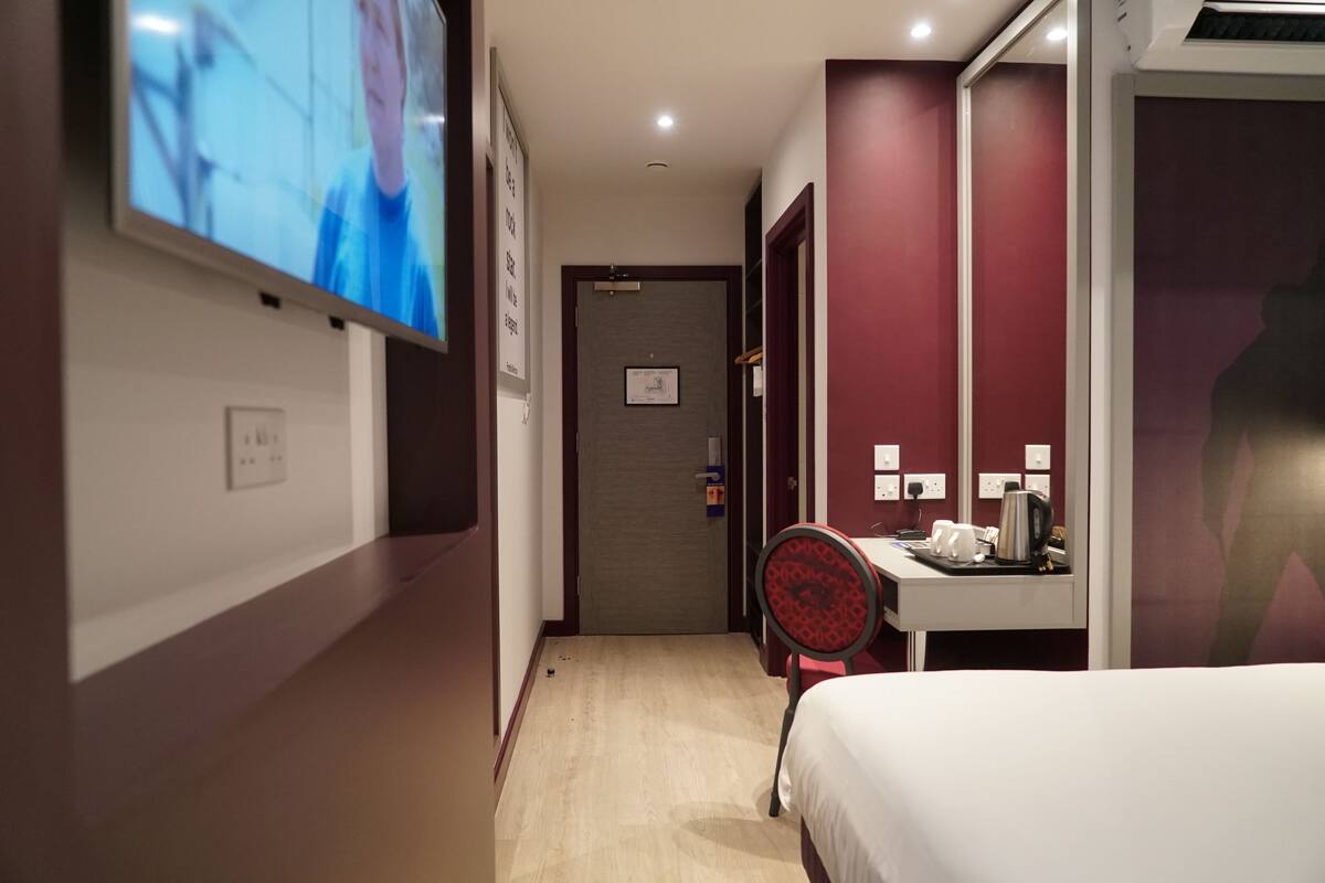 A hotel room with a bed, a desk with a chair, a TV mounted on the wall, and a door leading to another room.