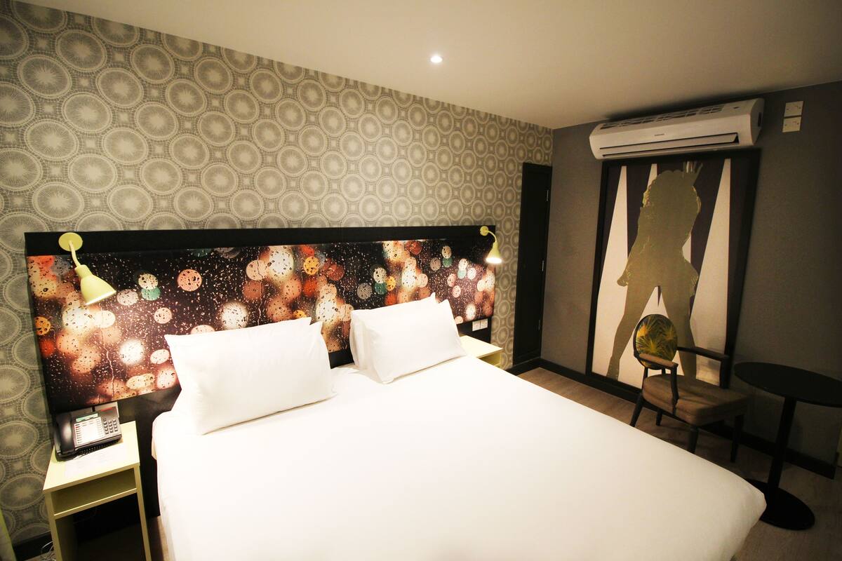 A hotel room with a patterned wallpaper, a bed with white linens, a nightstand with a phone, and an air conditioning unit.