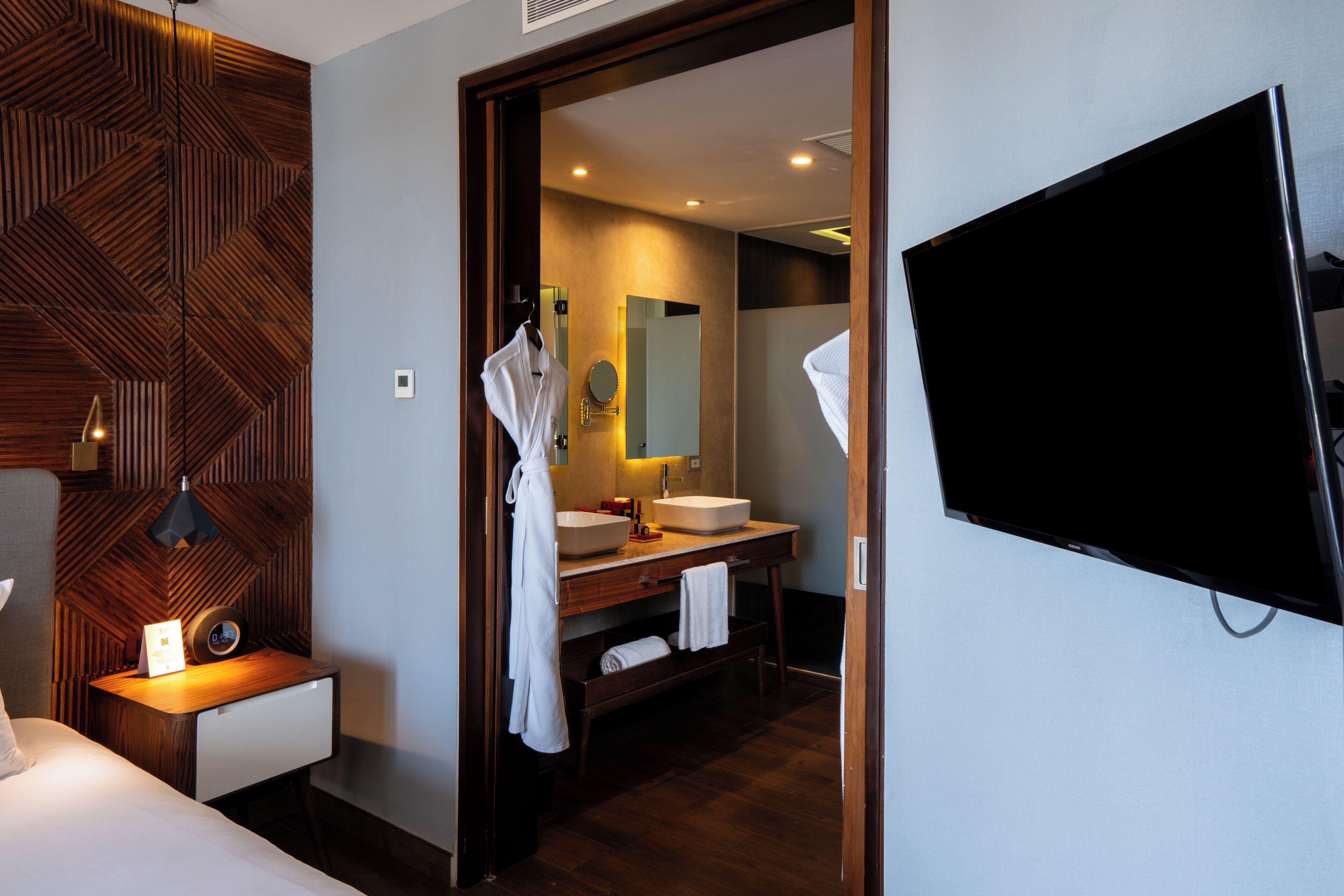 Une chambre d’hôtel avec un mur d’accent en bois, une table de chevet avec une lampe, un lit, une télévision, une salle de bain avec un lavabo et un miroir, et un peignoir accroché à la porte.