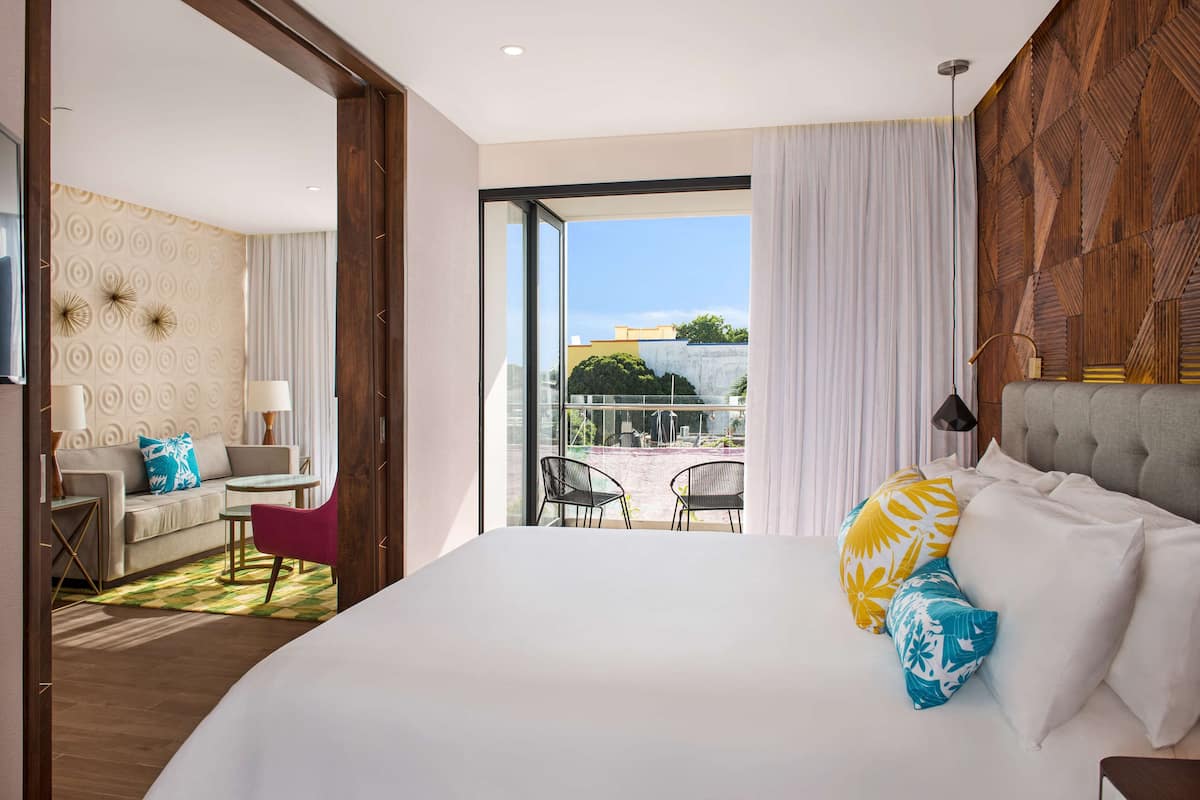 Ein Hotelzimmer mit einem großen Bett, einer Couch, einem kleinen Tisch und zwei Stühlen. Durch das Fenster ist ein Blick ins Freie.