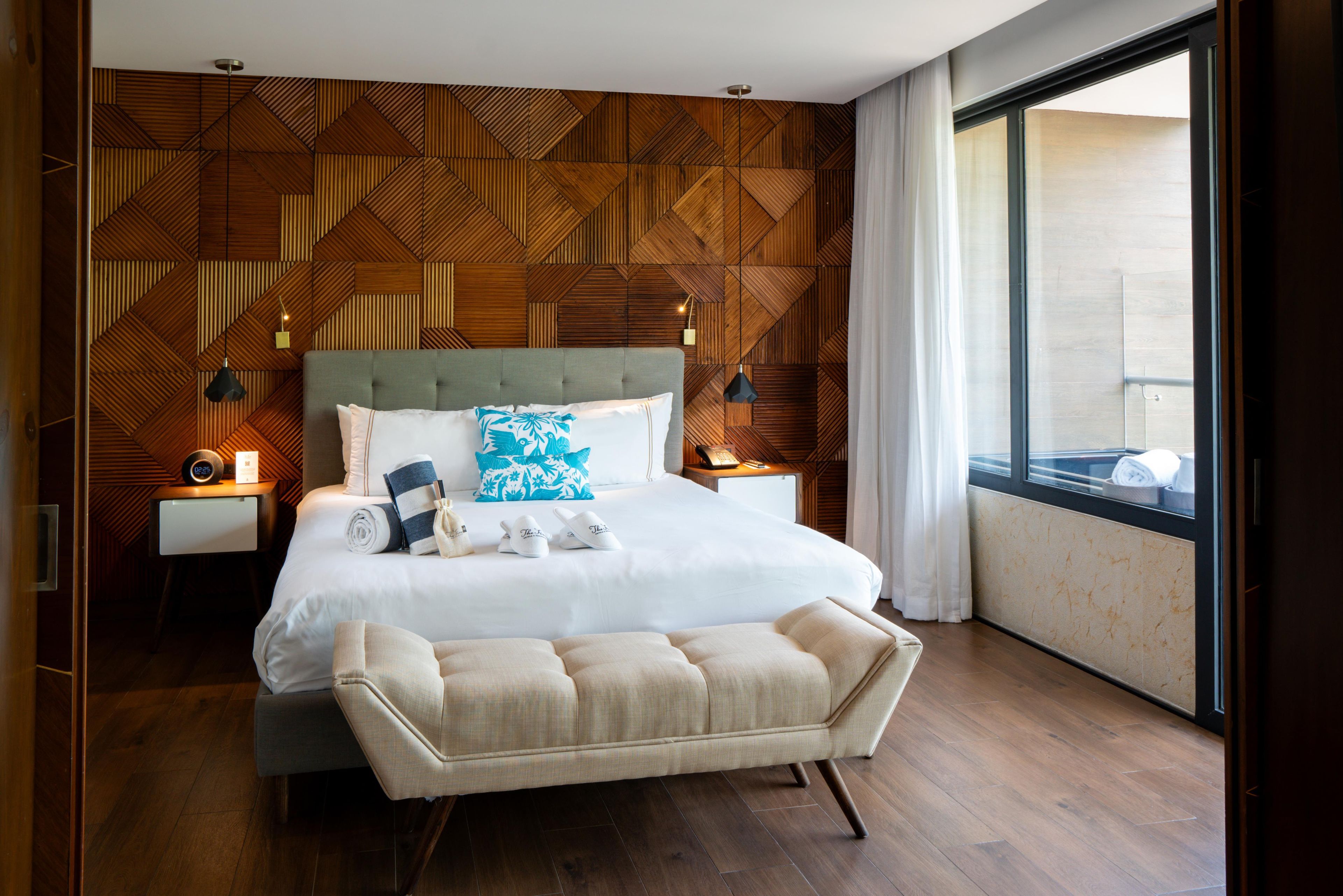 A modern hotel room with a geometric wooden wall design, a large bed with a tufted headboard, a bedside table with a lamp, and a view of the city through a large window.