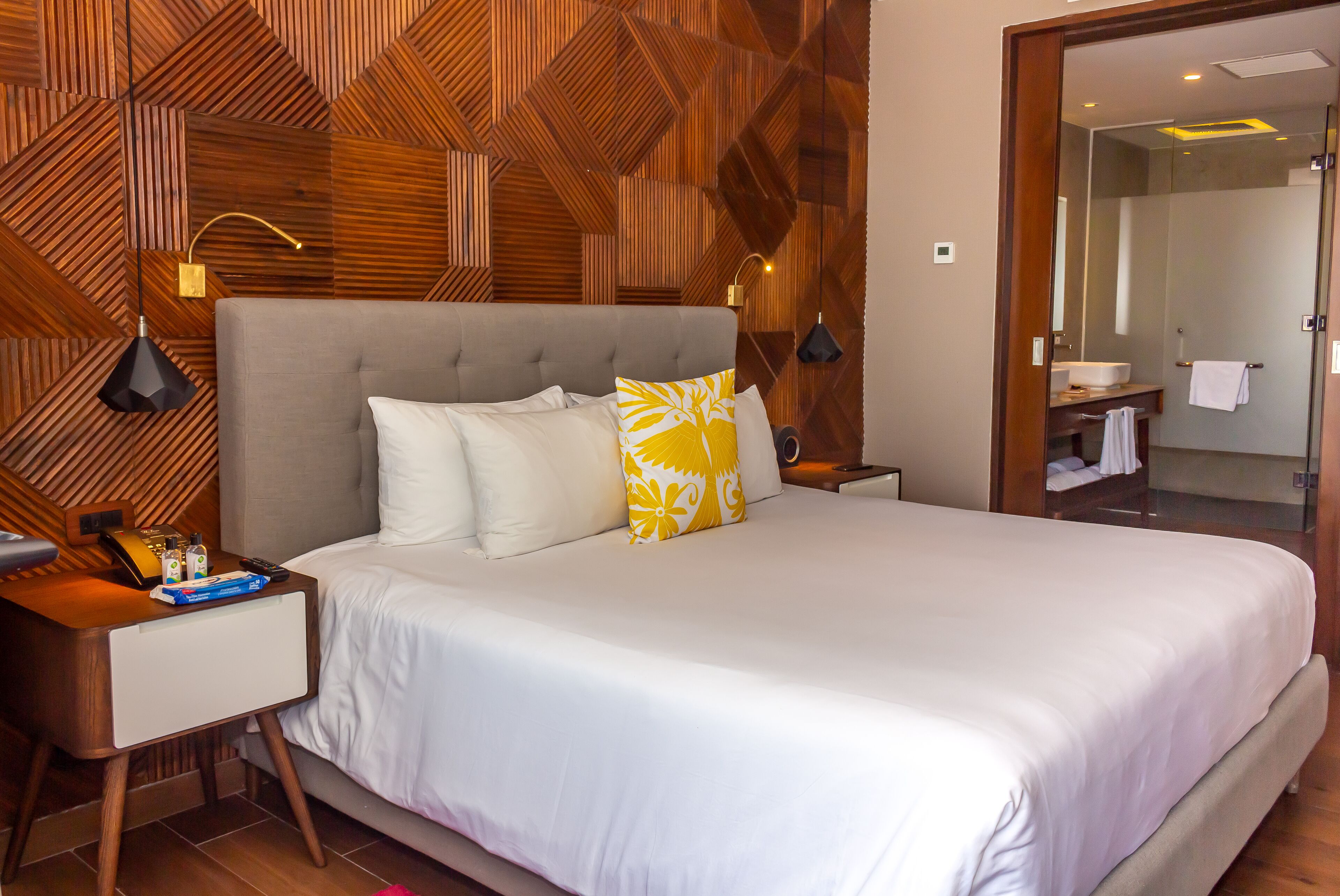 A modern hotel room with a large bed, wooden wall paneling, a bedside table with a lamp, and a bathroom visible through an open door.
