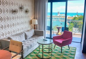 A modern living room with a sofa, a round table, and a chair, a large window with a view of the sea and buildings, and a patterned wall.
