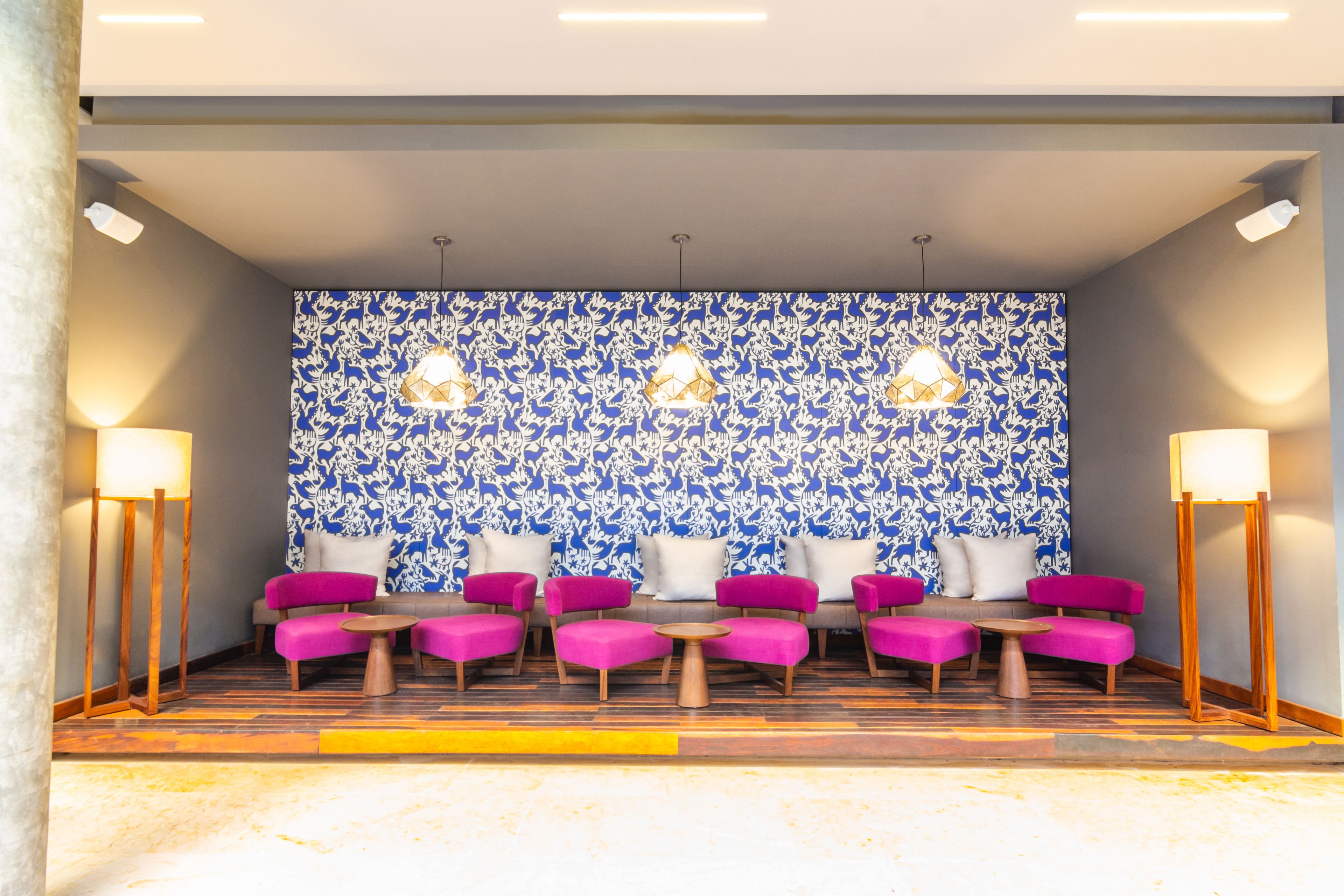 Un hall d’entrée avec un mur orné de motifs, plusieurs espaces de détente avec des fauteuils roses et blancs, et deux lampadaires.