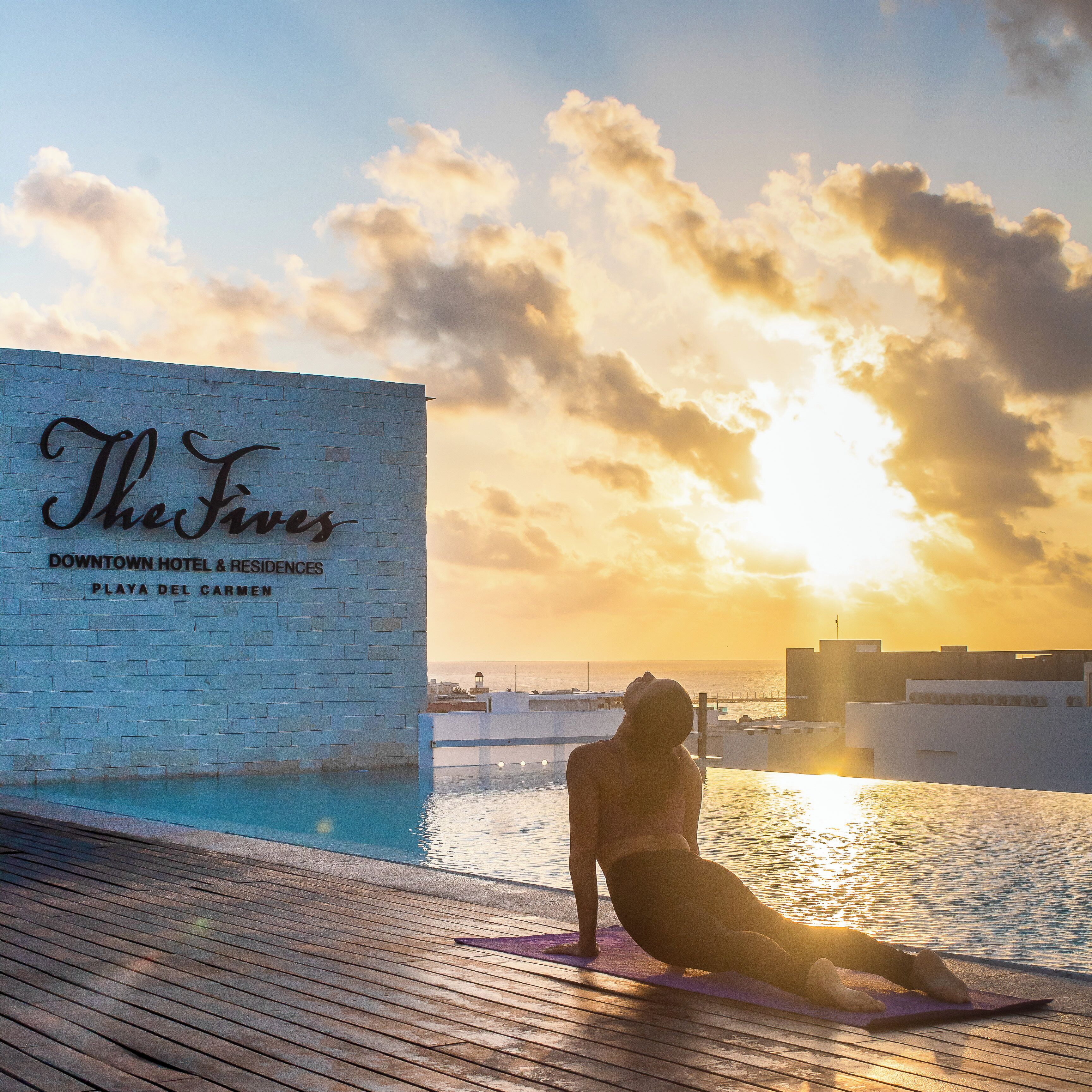 Una persona haciendo yoga en una terraza de madera con vista al océano y un edificio con un cartel que dice "The Fives Downtown Hotel & Residences Playa Del Carmen".