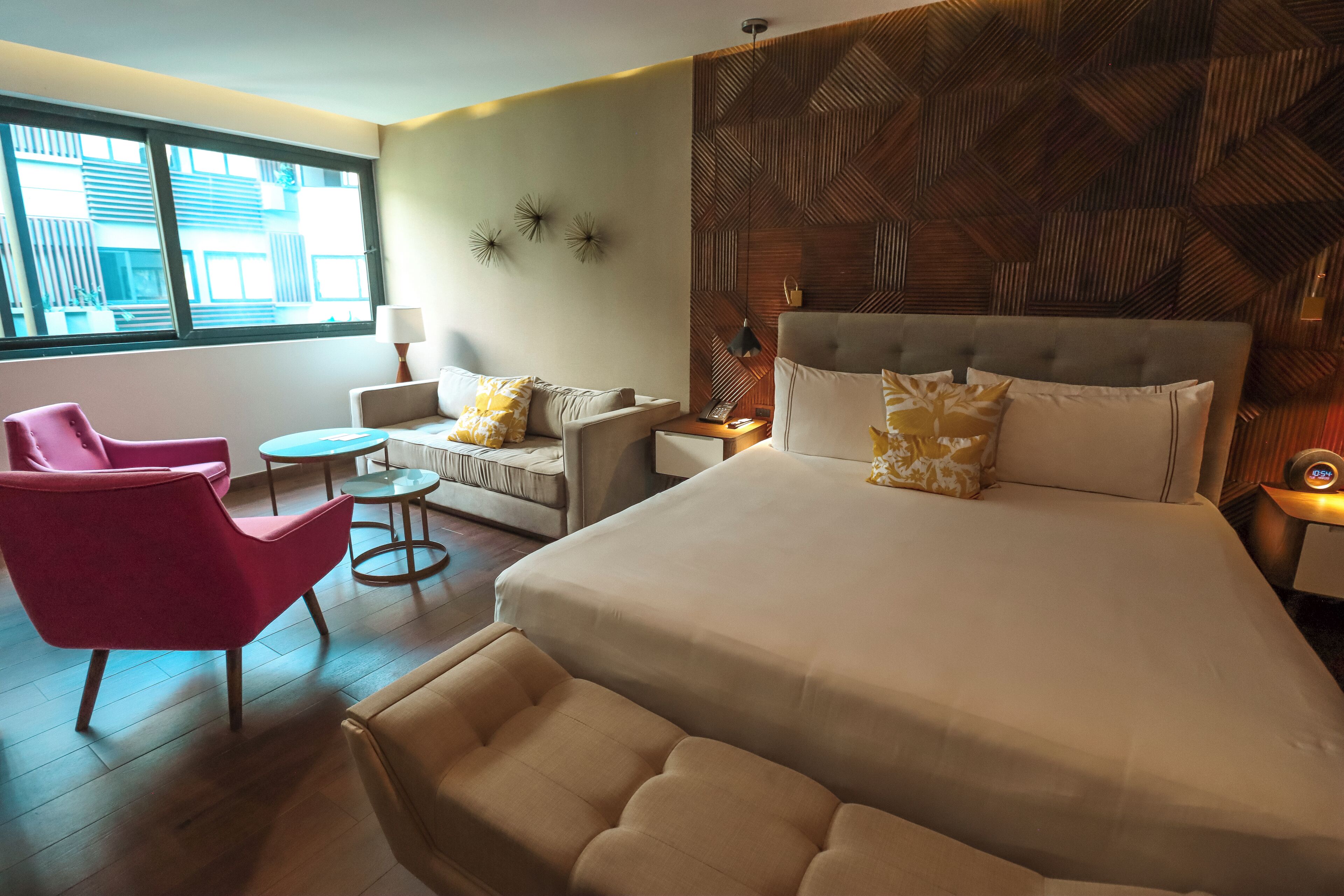 A modern hotel room with a large bed, a pink armchair, a small round table, and a sofa with patterned cushions.