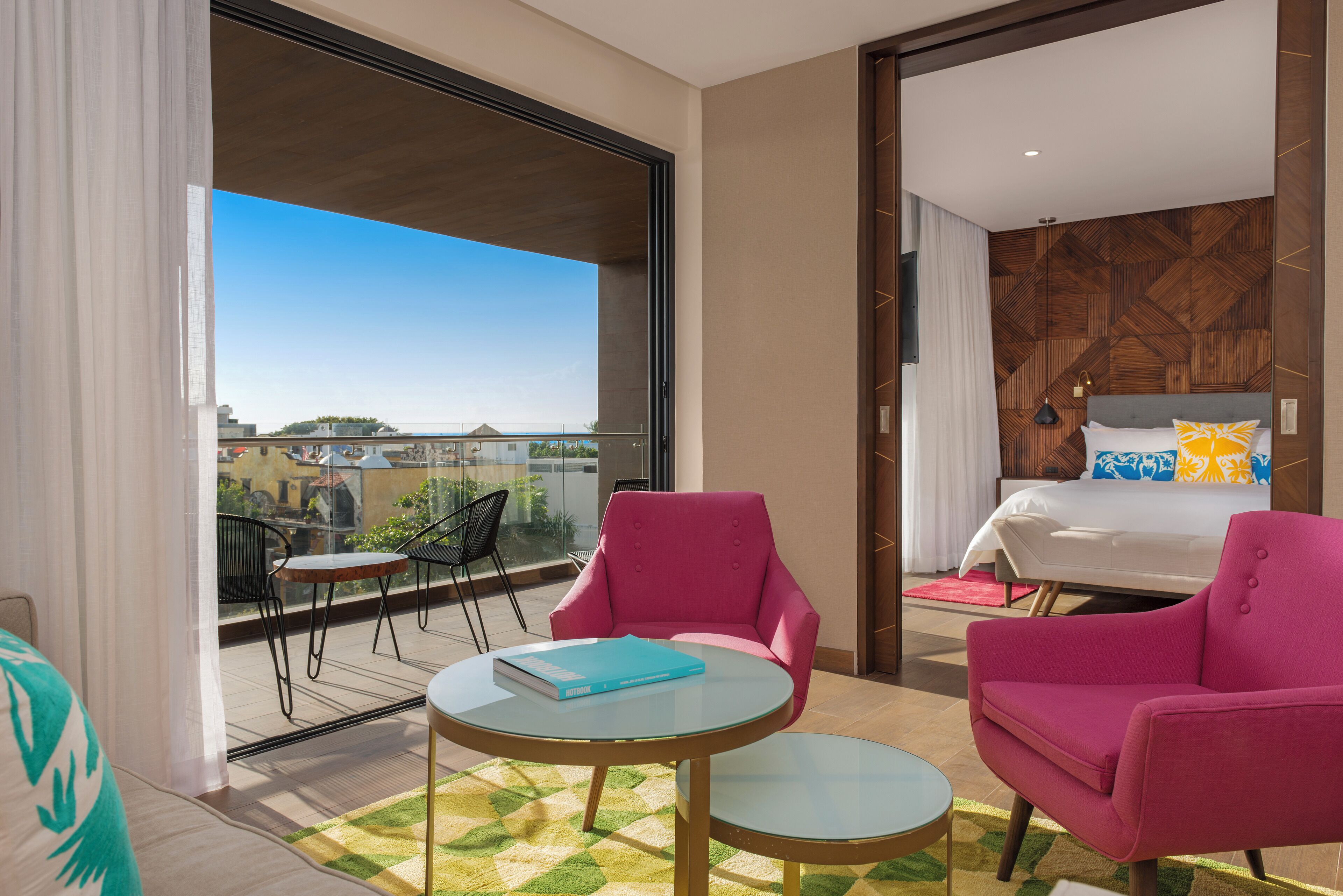 A hotel room with a balcony, a bed with a blue and white patterned headboard, and two pink armchairs.