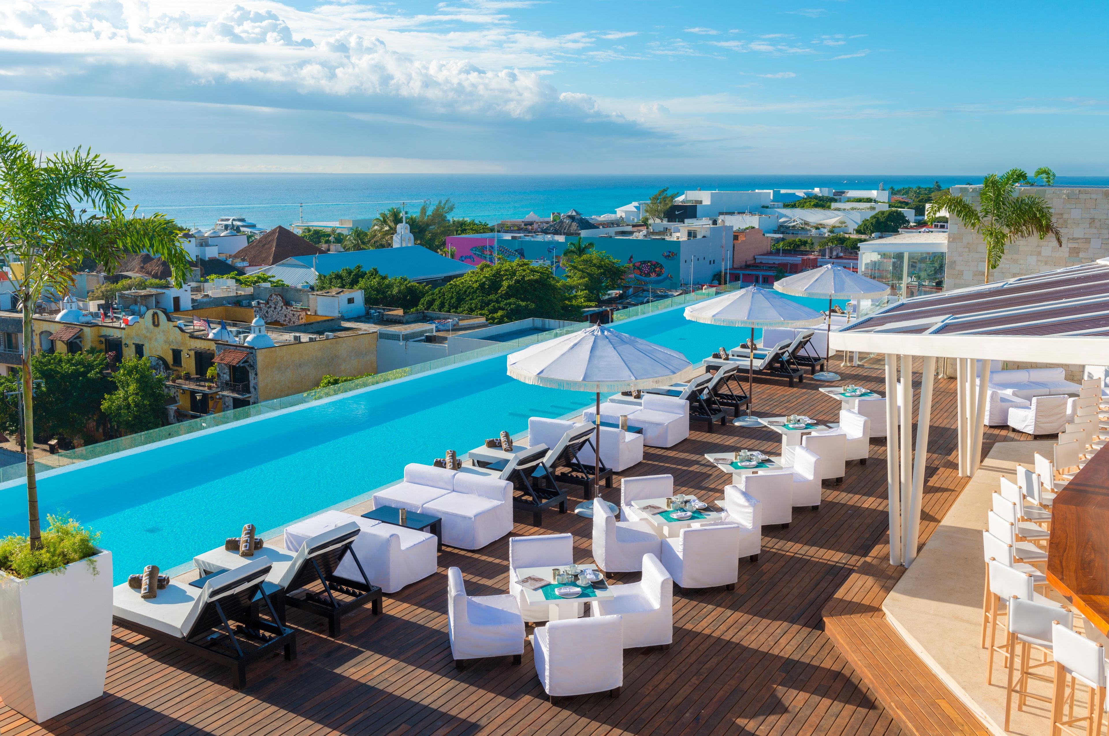 Un área de piscina en la azotea con sillones blancos y sombrillas, con vista al mar azul turquesa y un pueblo costero.