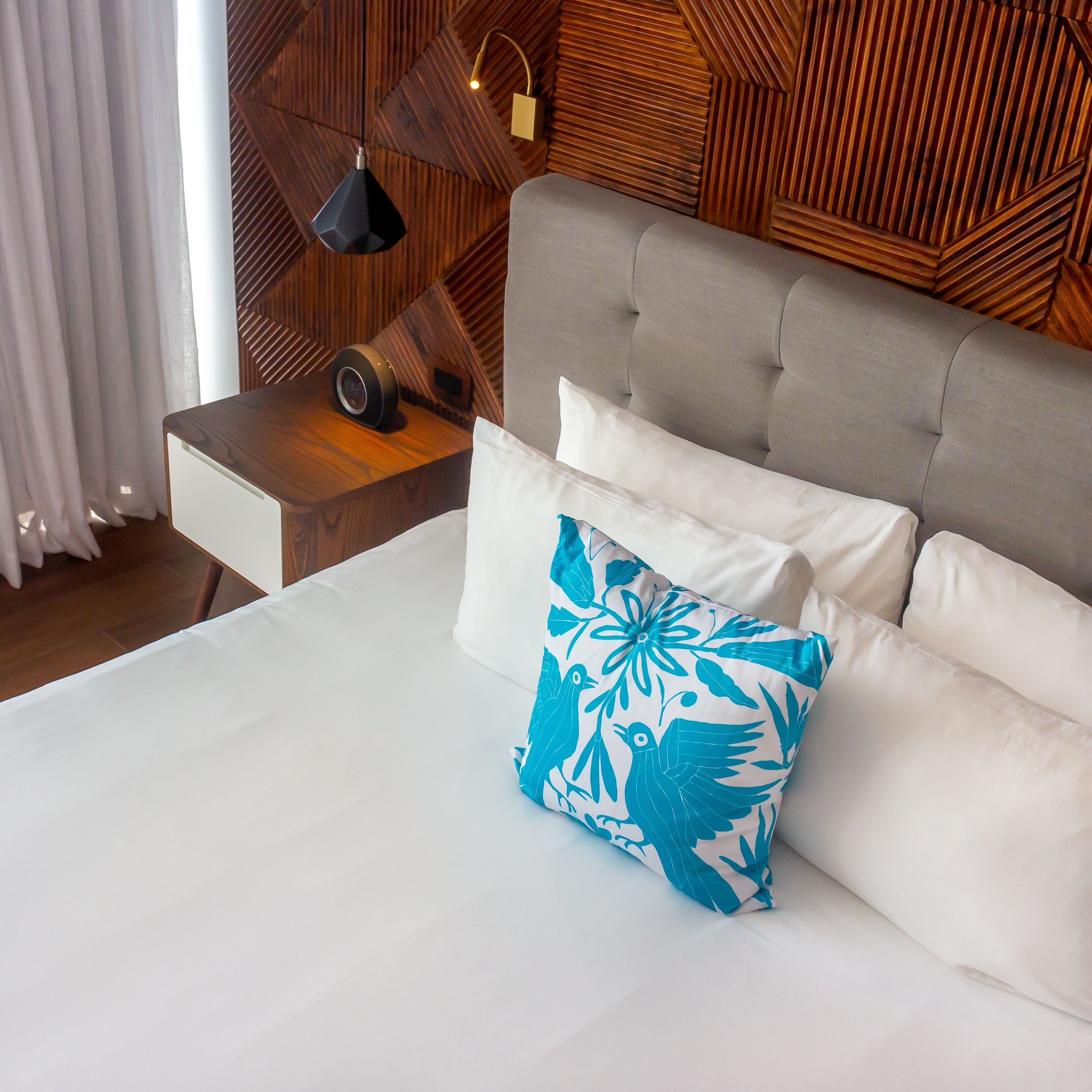 Una cama bien tendida con almohadones blancos y azules estampados, una mesita de noche de madera con un reloj y una pared con diseño geométrico de paneles de madera.