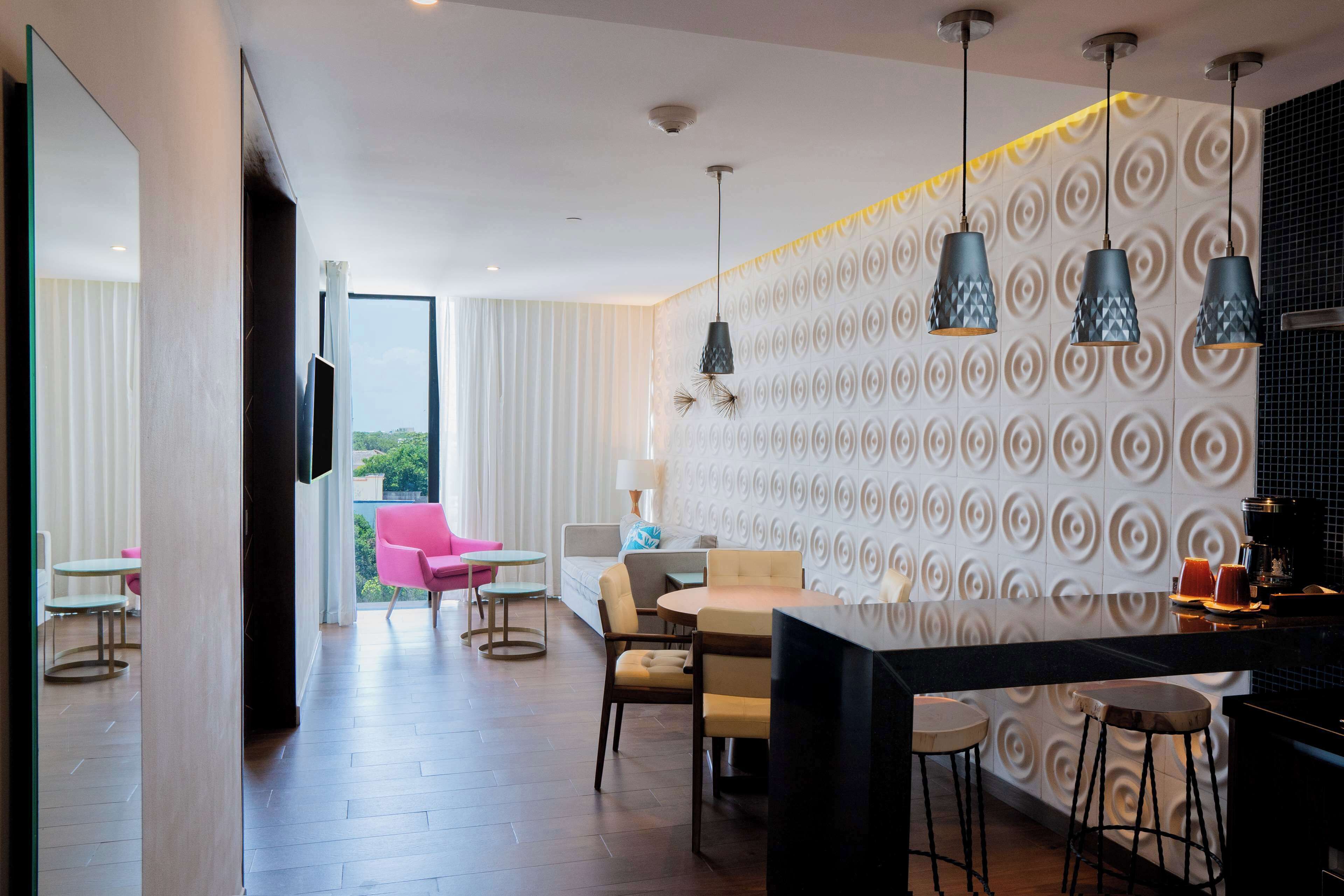 A modern hotel room with a dining area, a kitchen, and a balcony with pink chairs.