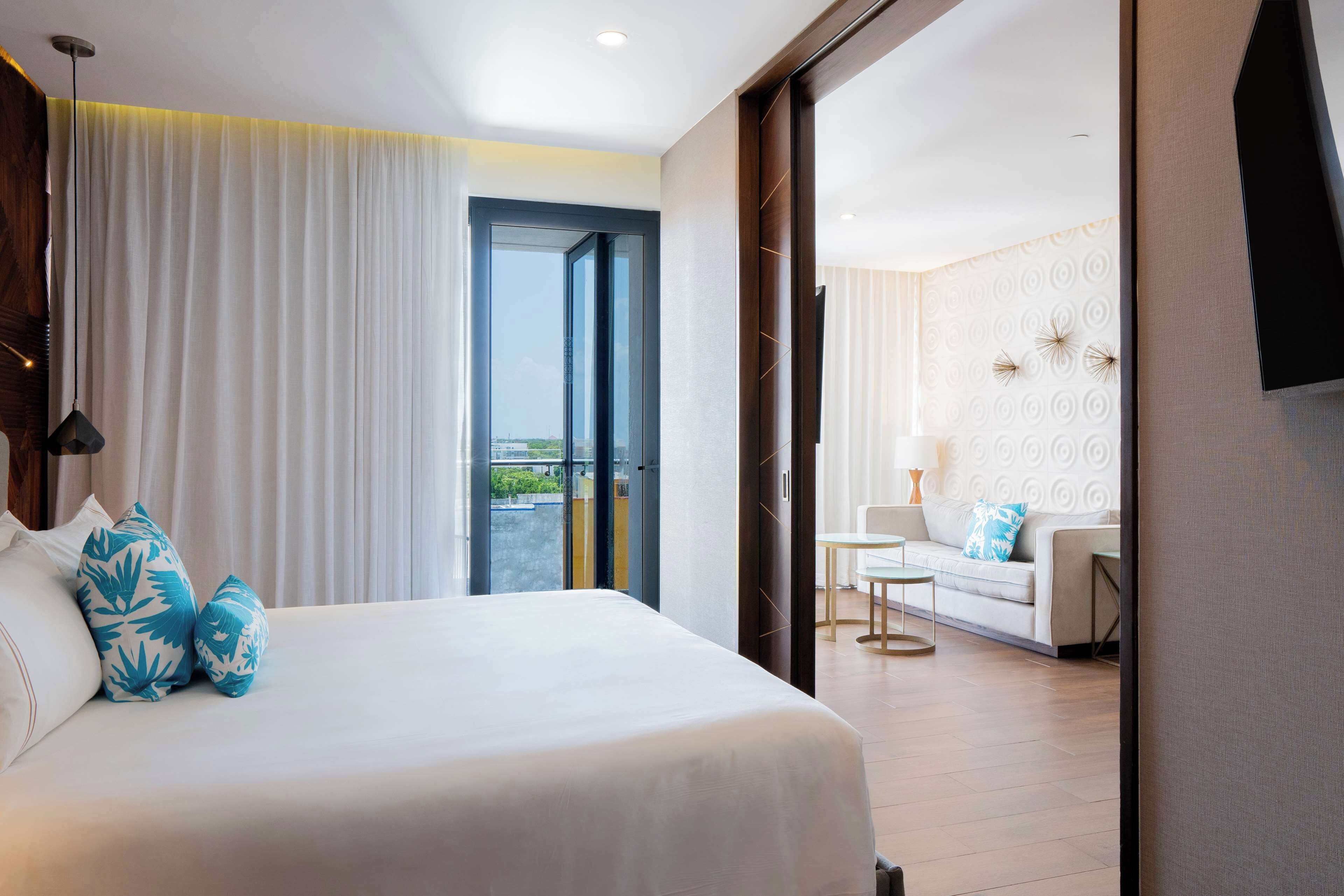 Quarto de hotel com uma cama grande, varanda com vista, área de estar com sofá e mesa, e televisão na parede.