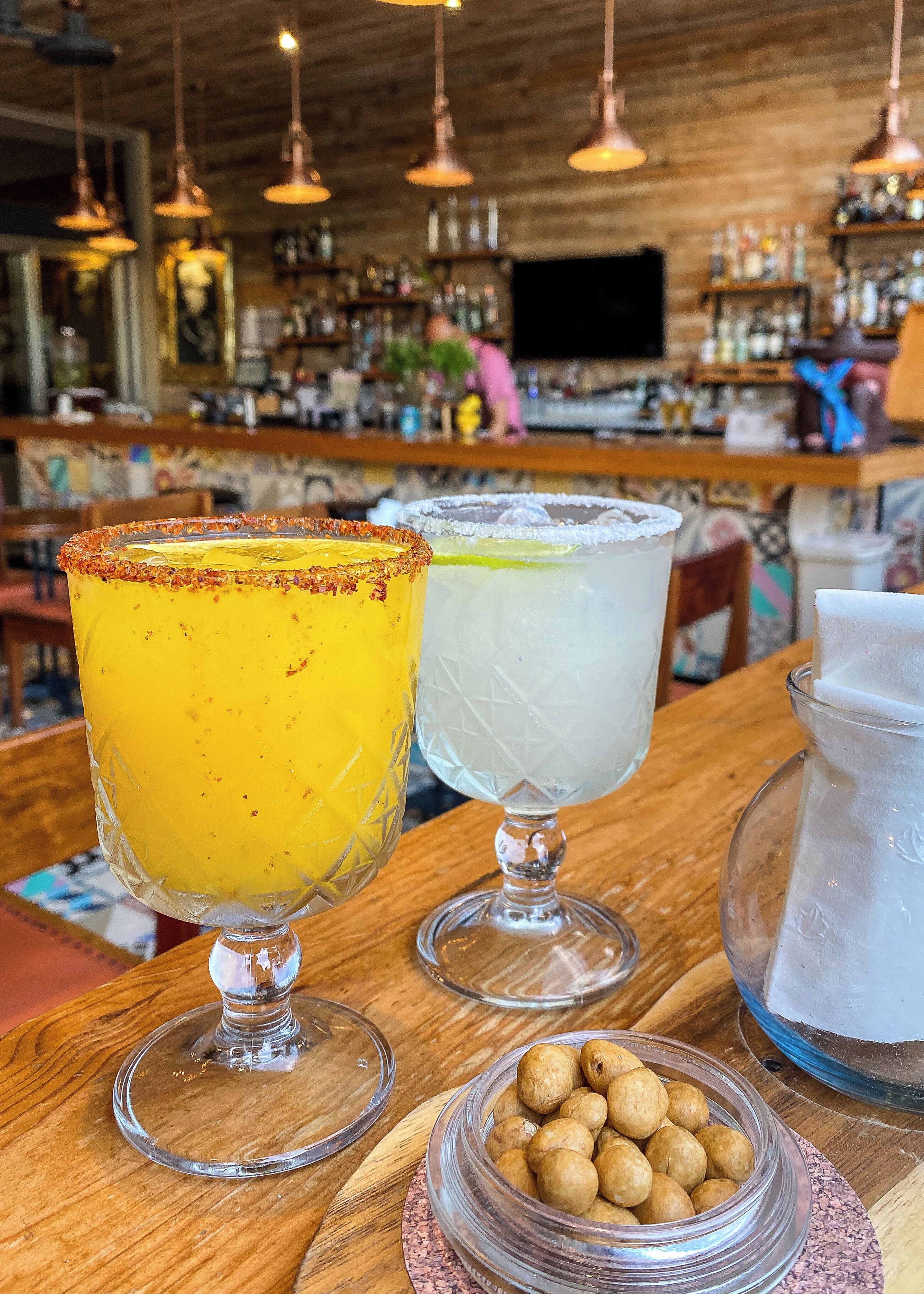 Dos margaritas sobre una barra de madera con una jarra de hielo y un tazón de nueces.