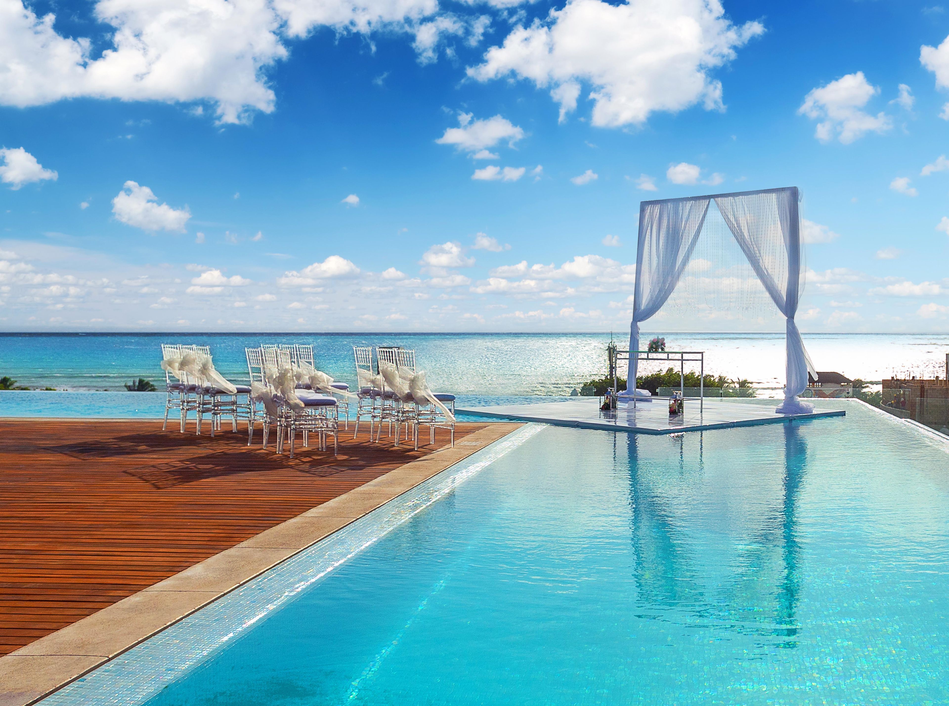 Piscina infinita com deck de madeira e cadeiras brancas, com vista para uma praia.