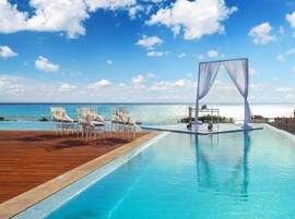 Piscina infinita com deck de madeira e cadeiras brancas, com vista para uma praia.