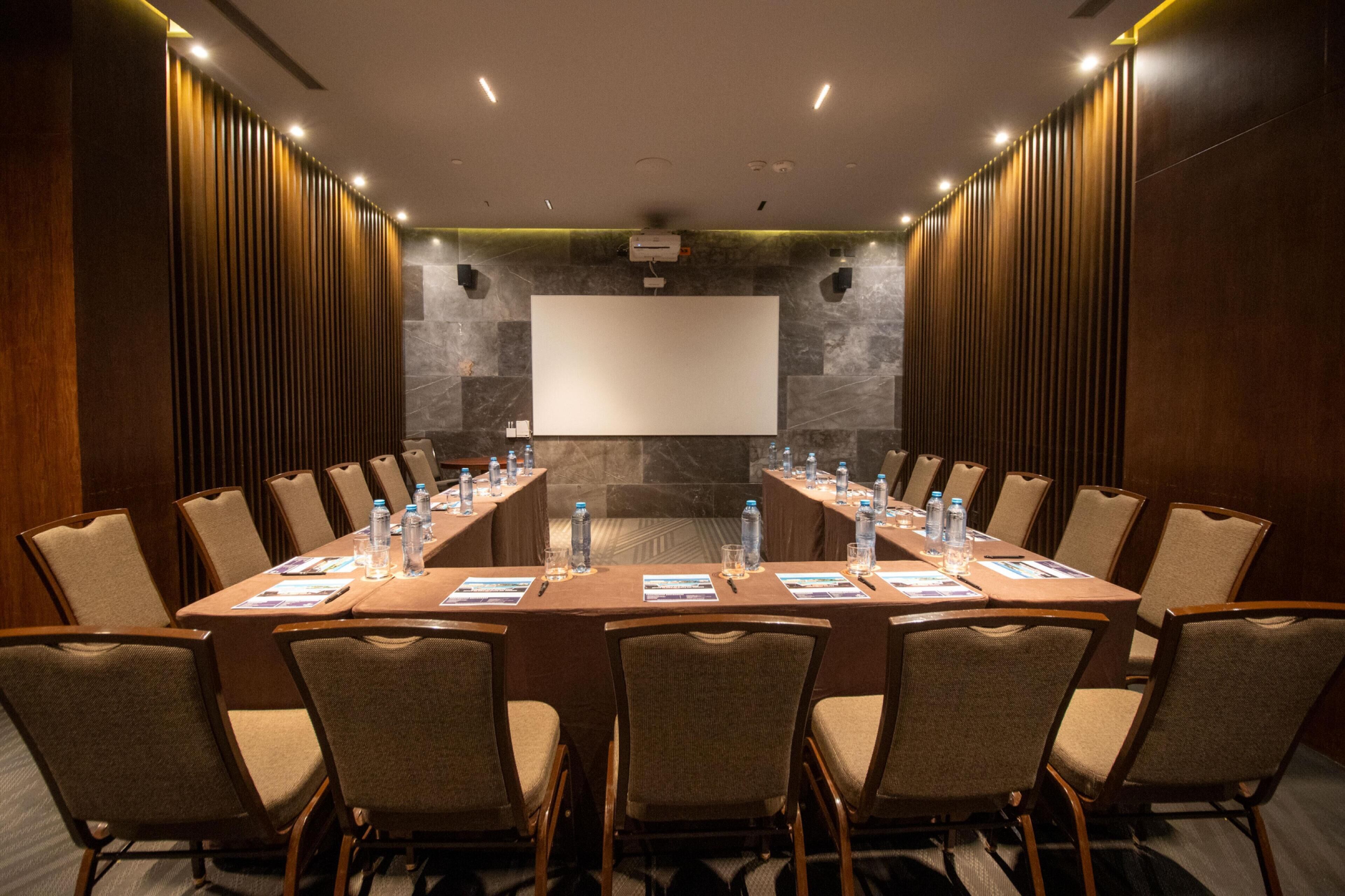 A conference room with a long table, chairs, a projector screen, and a stone wall.