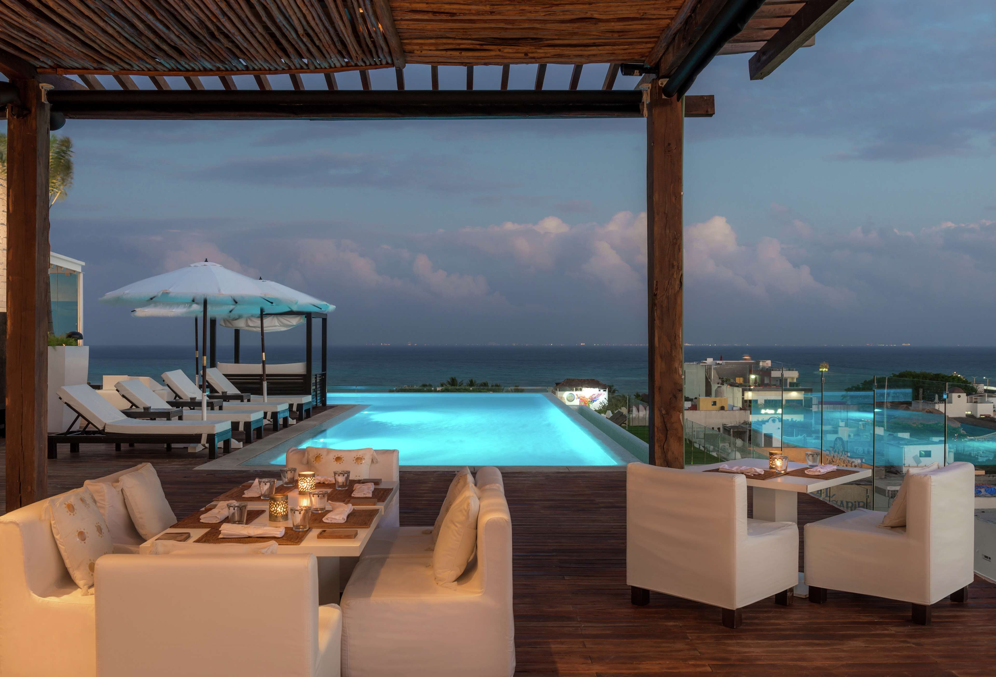 A poolside area with lounge chairs, a dining table, and a view of the ocean at dusk.