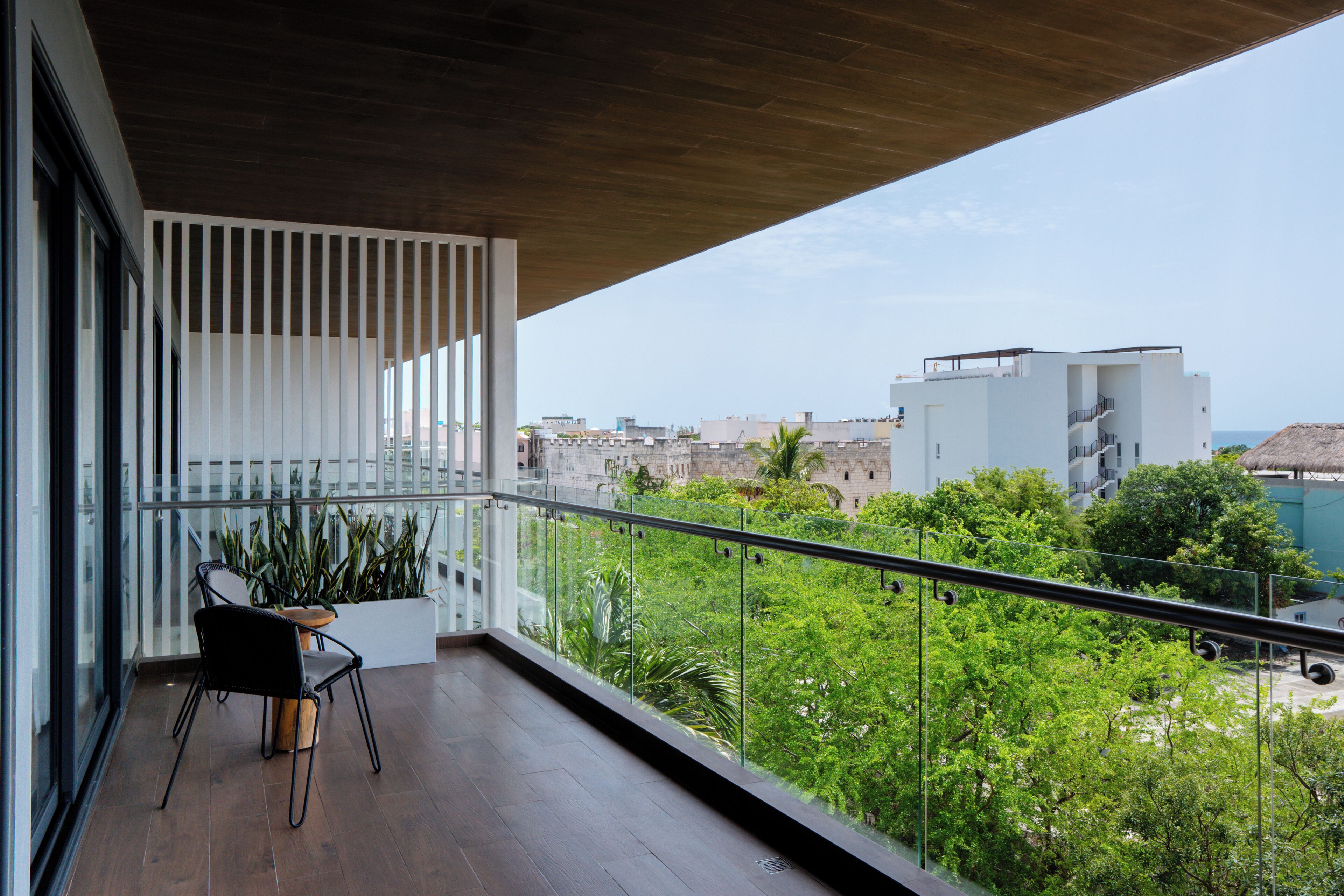 Un balcón con piso de madera, baranda de vidrio y vista a un paisaje urbano y zonas verdes.