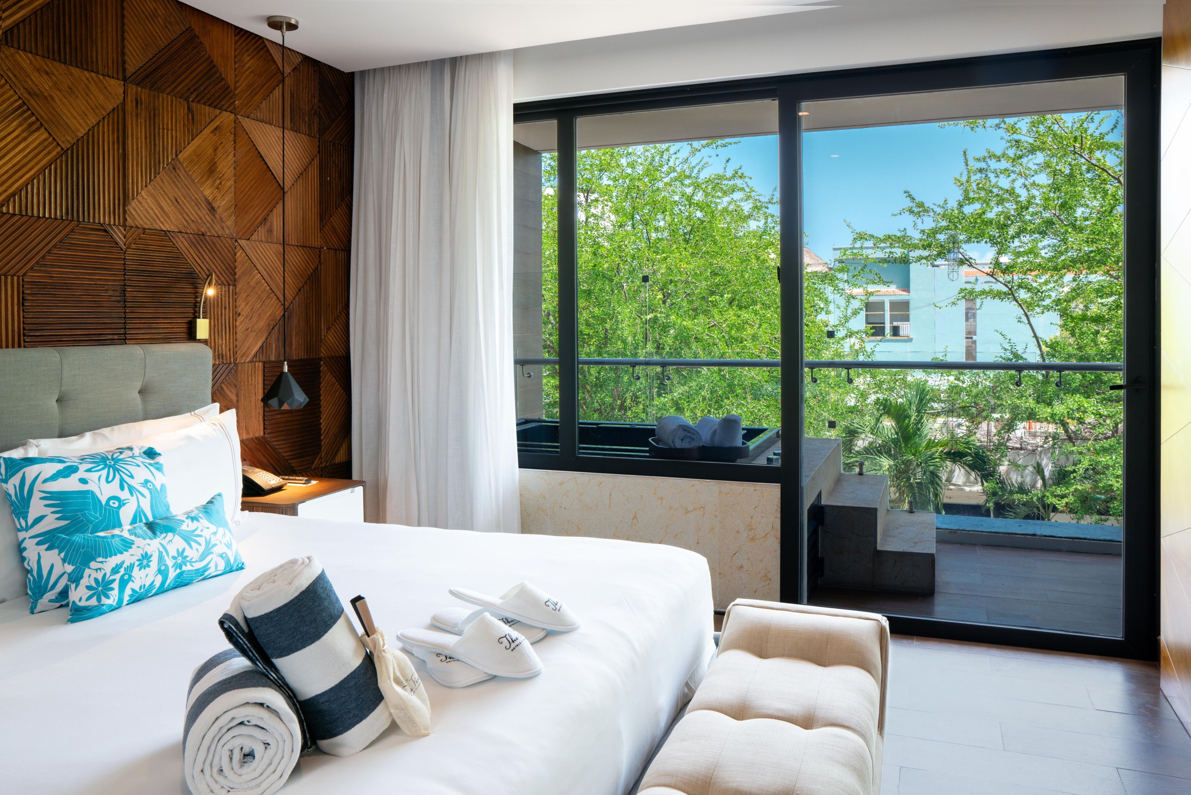 Una habitación de hotel moderna con una cama grande, vistas a zonas verdes y edificios, y un balcón con zona de estar.
