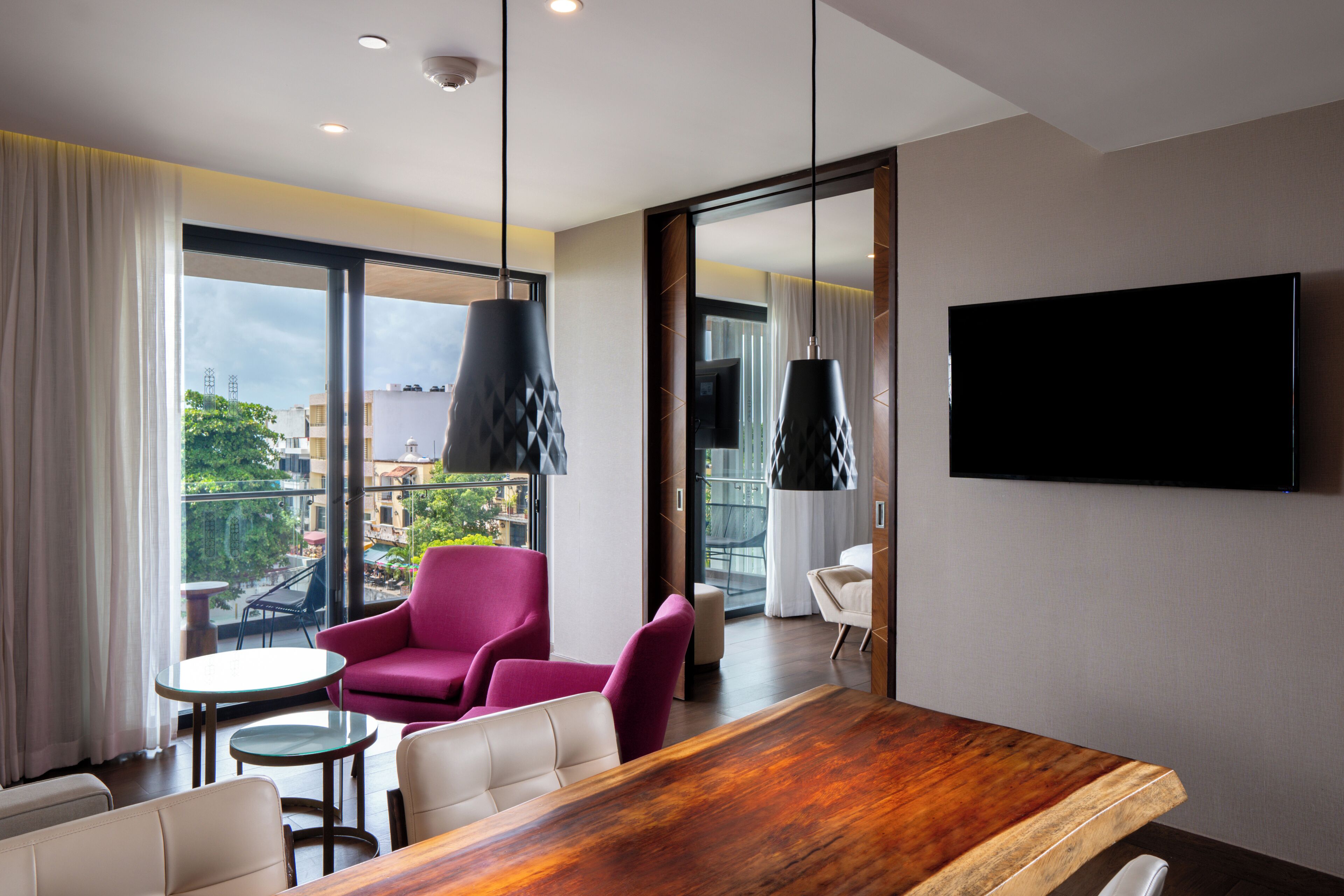Une chambre d’hôtel moderne avec une table à manger en bois, deux chaises, un fauteuil rose et un téléviseur fixé au mur.