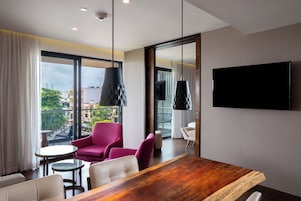 Una habitación de hotel moderna con una mesa de comedor de madera, dos sillas, un sillón rosa y un televisor montado en la pared.