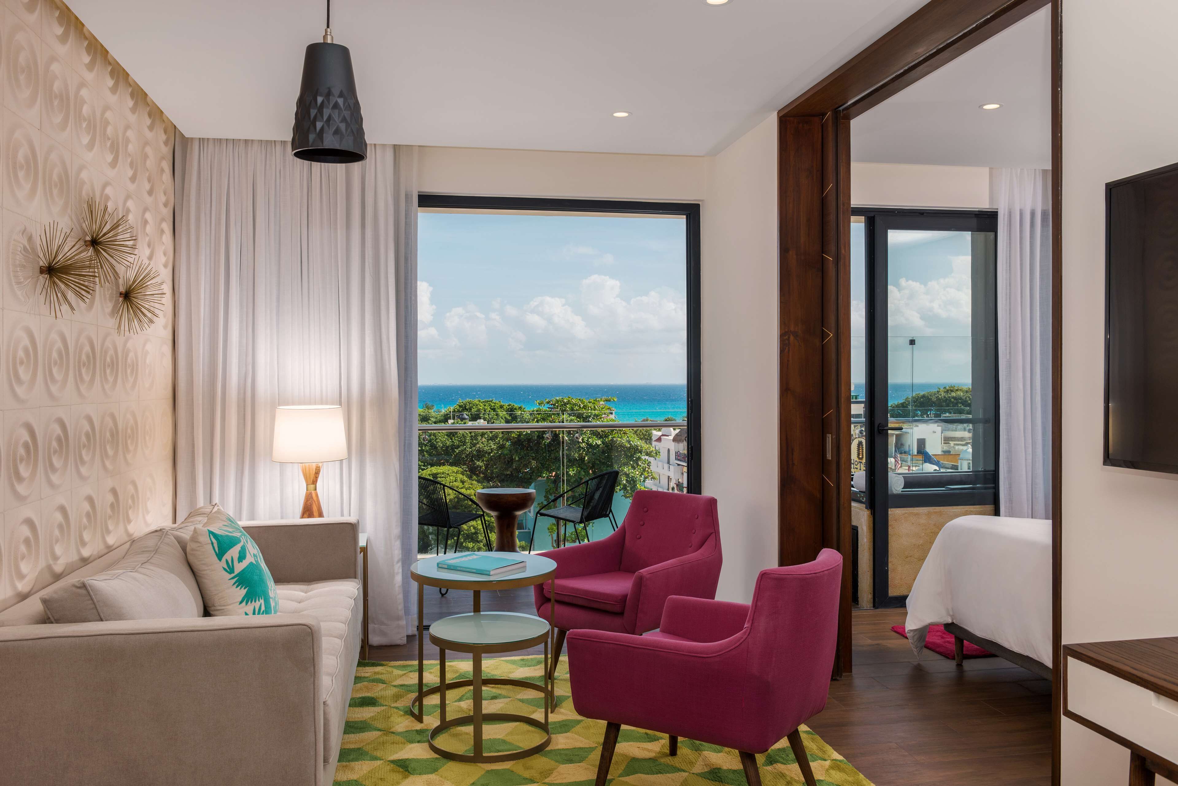 A modern hotel room with a balcony, a sofa, two pink chairs, a small round table, and a bed.