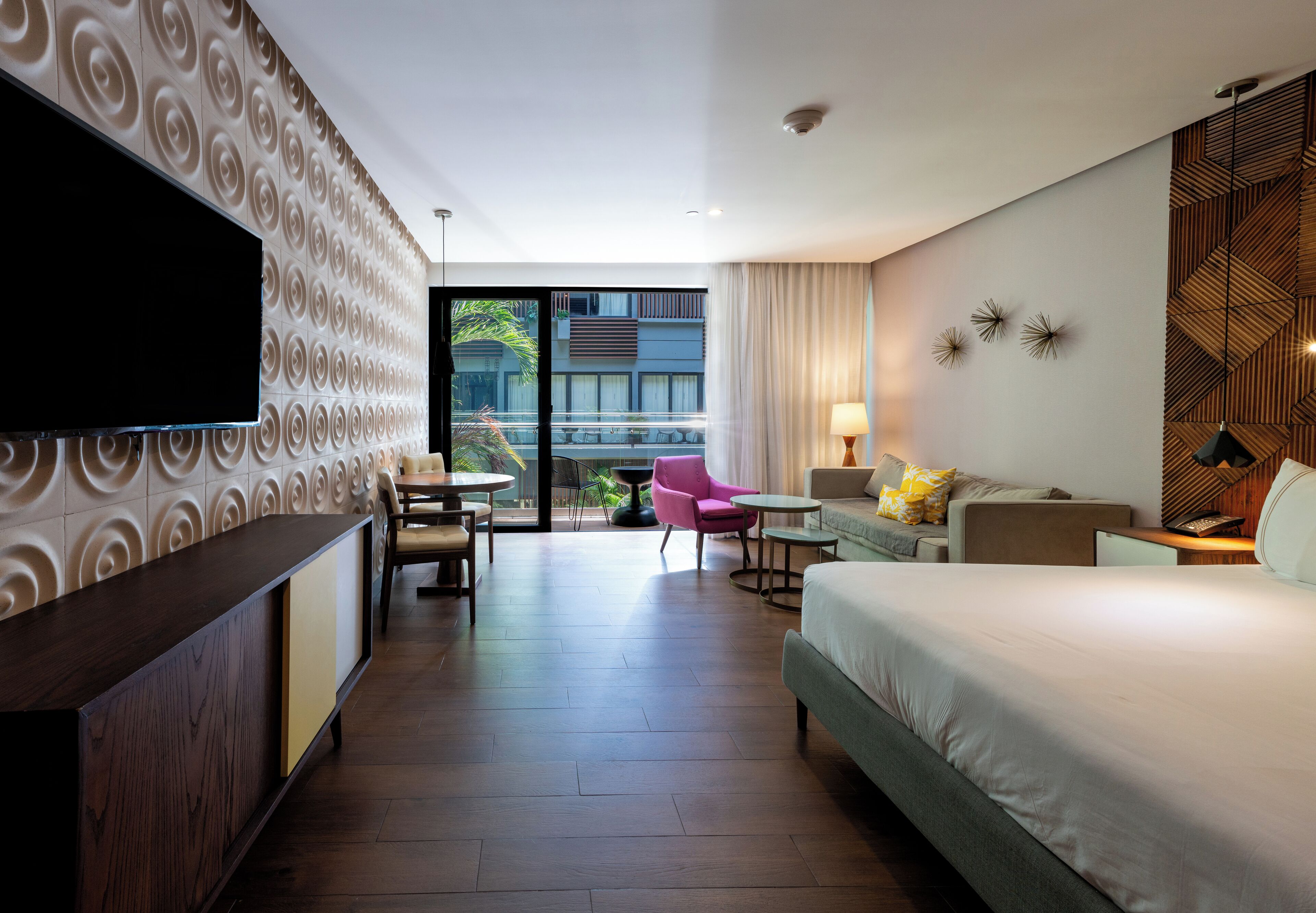 A hotel room with a large bed, a TV, a desk, a chair, and a sofa. There is a window with a view of the outdoors.