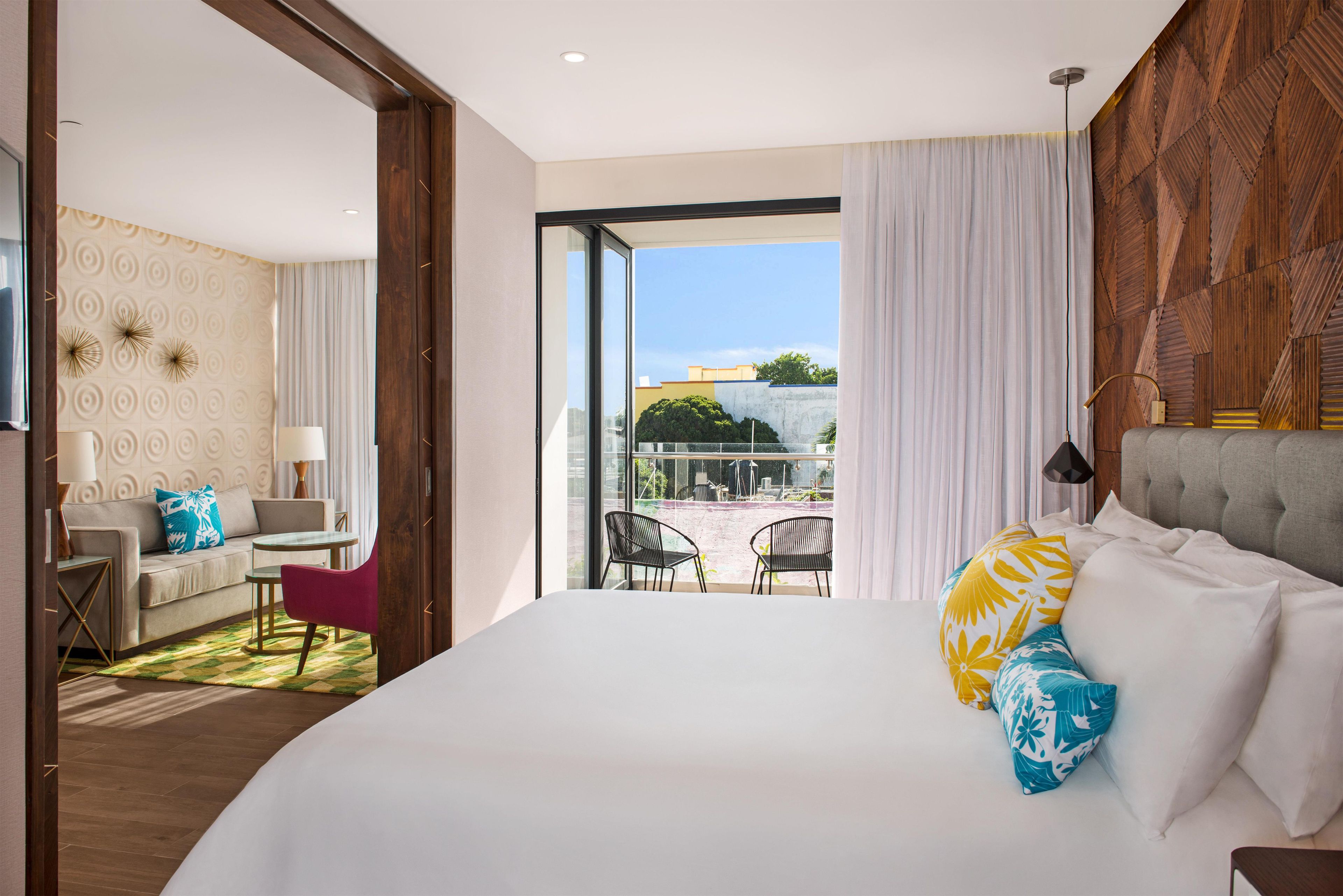 A hotel room with a large bed, a sofa, a dining table, and chairs. There is a balcony with outdoor seating.
