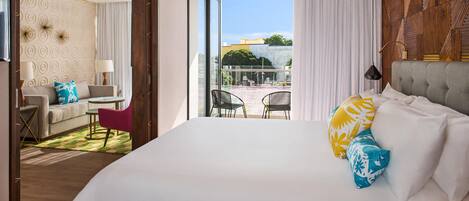 A hotel room with a large bed, a sofa, a dining table, and chairs. There is a balcony with outdoor seating.