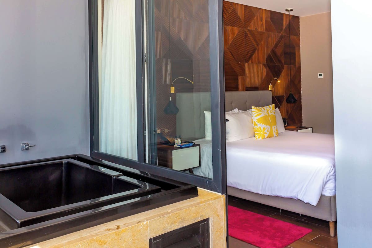 Ein modernes Hotelzimmer mit Duschkabine aus Glas, einem Bett mit weißer Bettwäsche und einem gelben Kissen sowie einem roten Teppich.