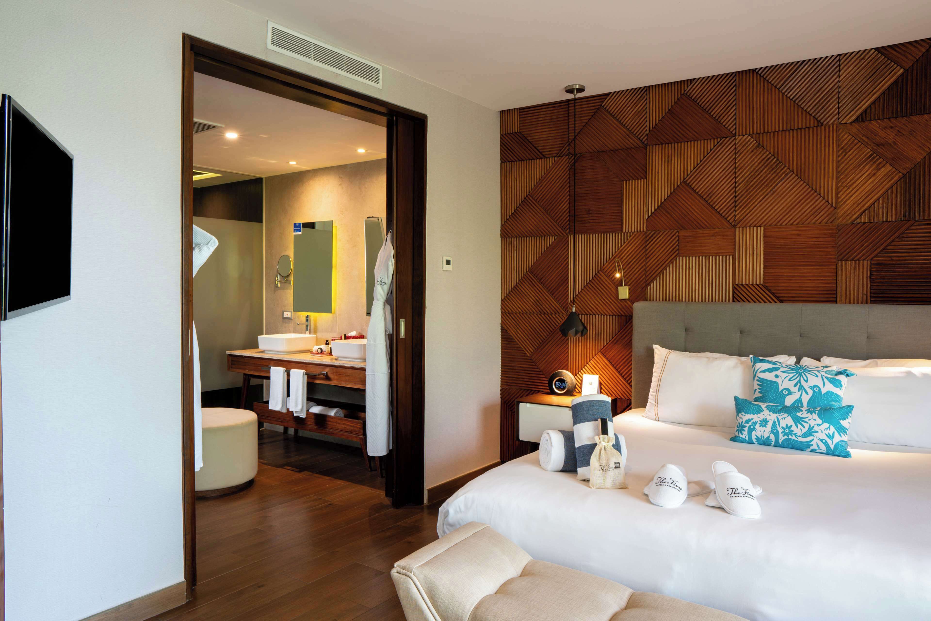 Una habitación de hotel moderna con una cama grande, un muro decorativo de madera, un baño con tocador y espejo, y un televisor de pantalla plana montado en la pared.