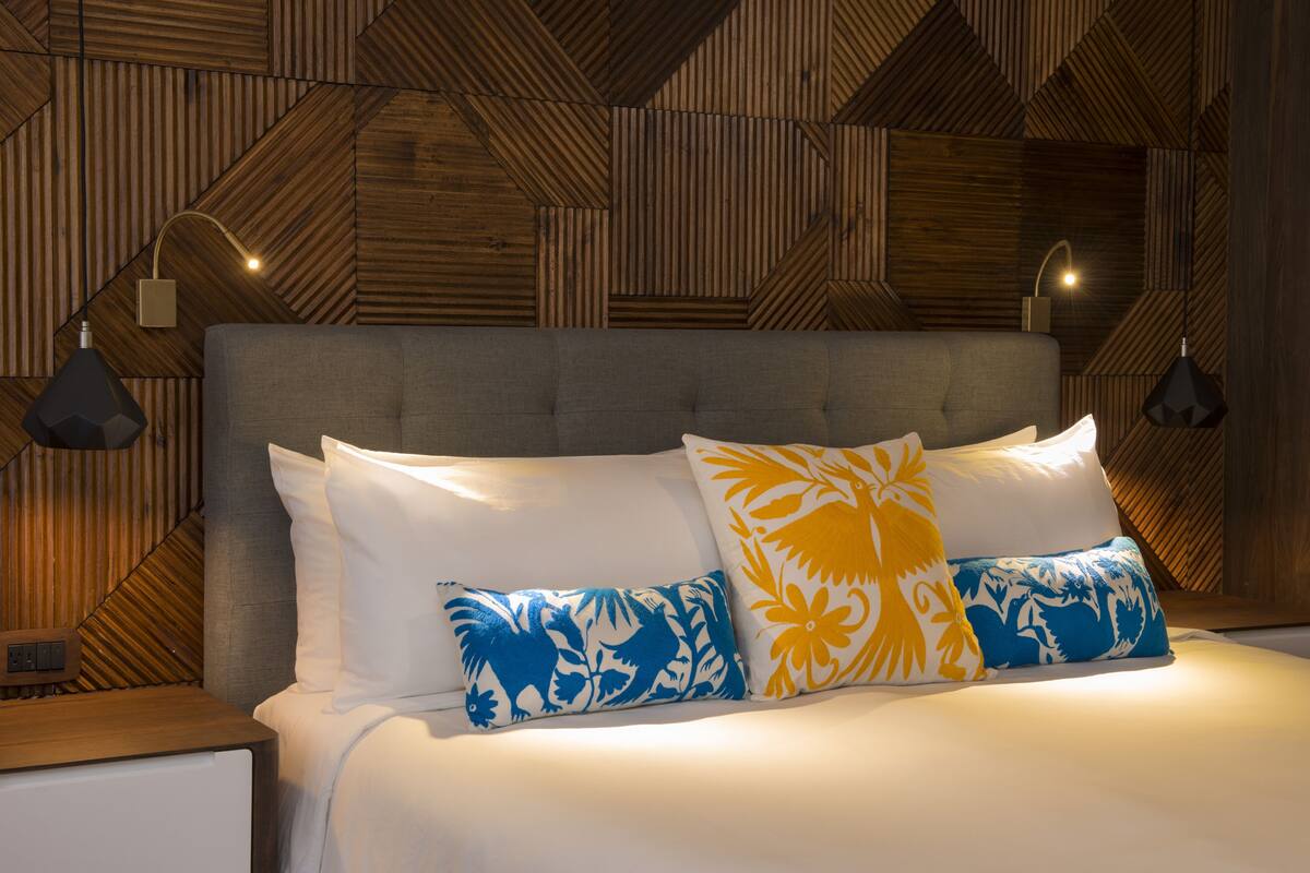 Ein Schlafzimmer mit einer Wand im Holzdekor mit geometrischen Mustern, einem grauen Stoffkopfende und zwei Kissen mit Muster.