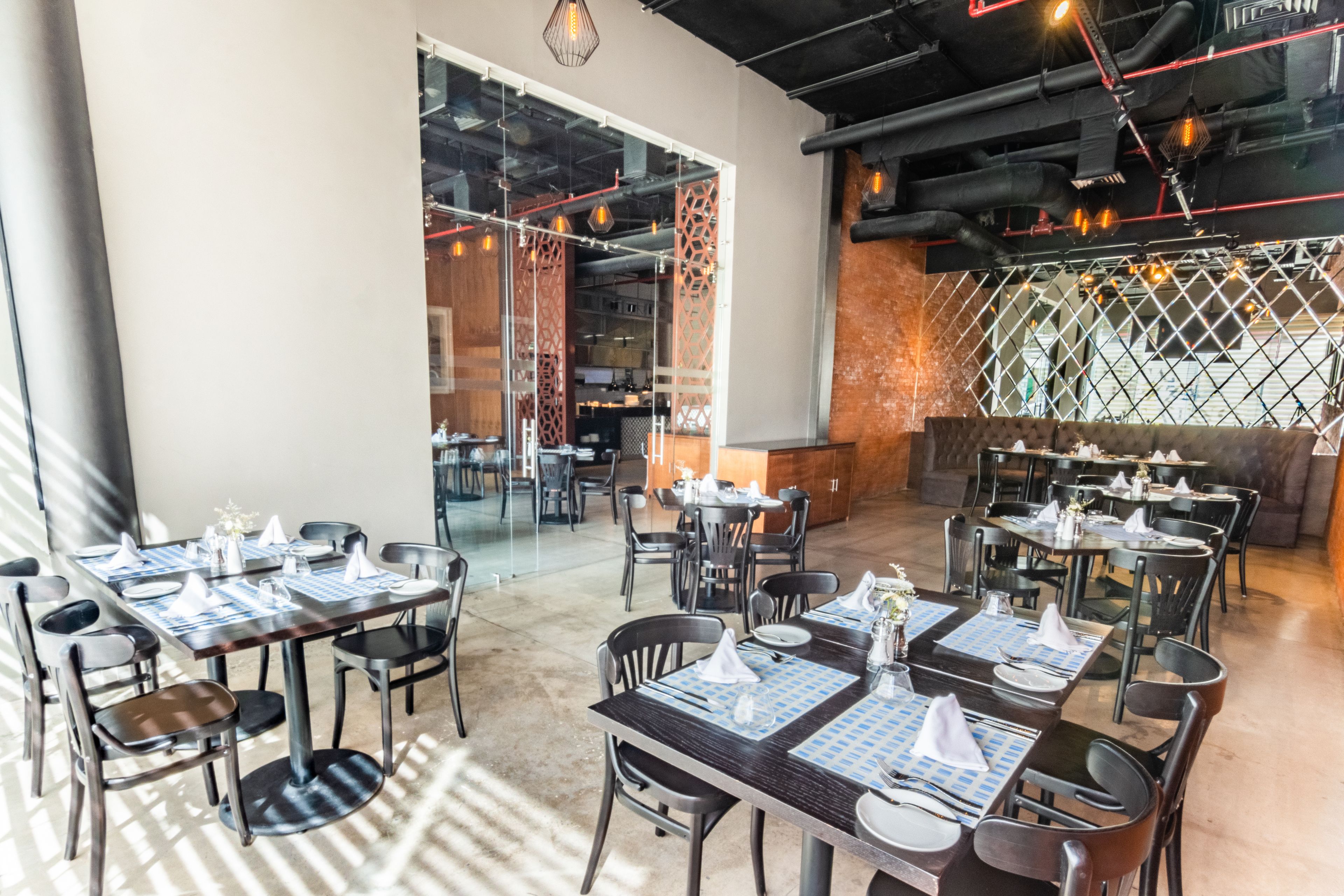 A modern restaurant with multiple dining tables and chairs, a glass partition separating the dining area from the kitchen, and a brick wall with a lattice design.